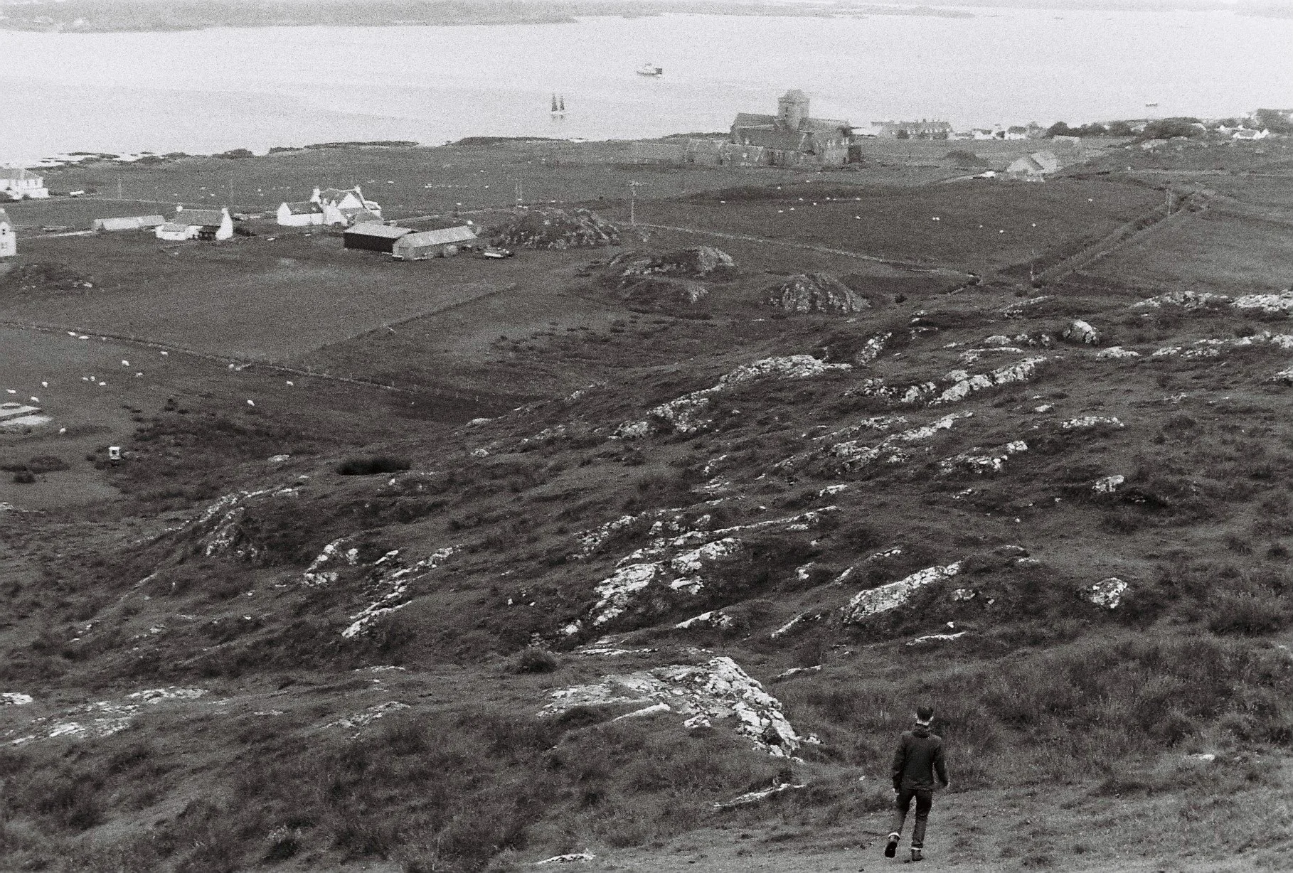A person walking down a hill toward a coastal landscape with rocky terrain, scattered houses, and a body of water with boats, in black and white.