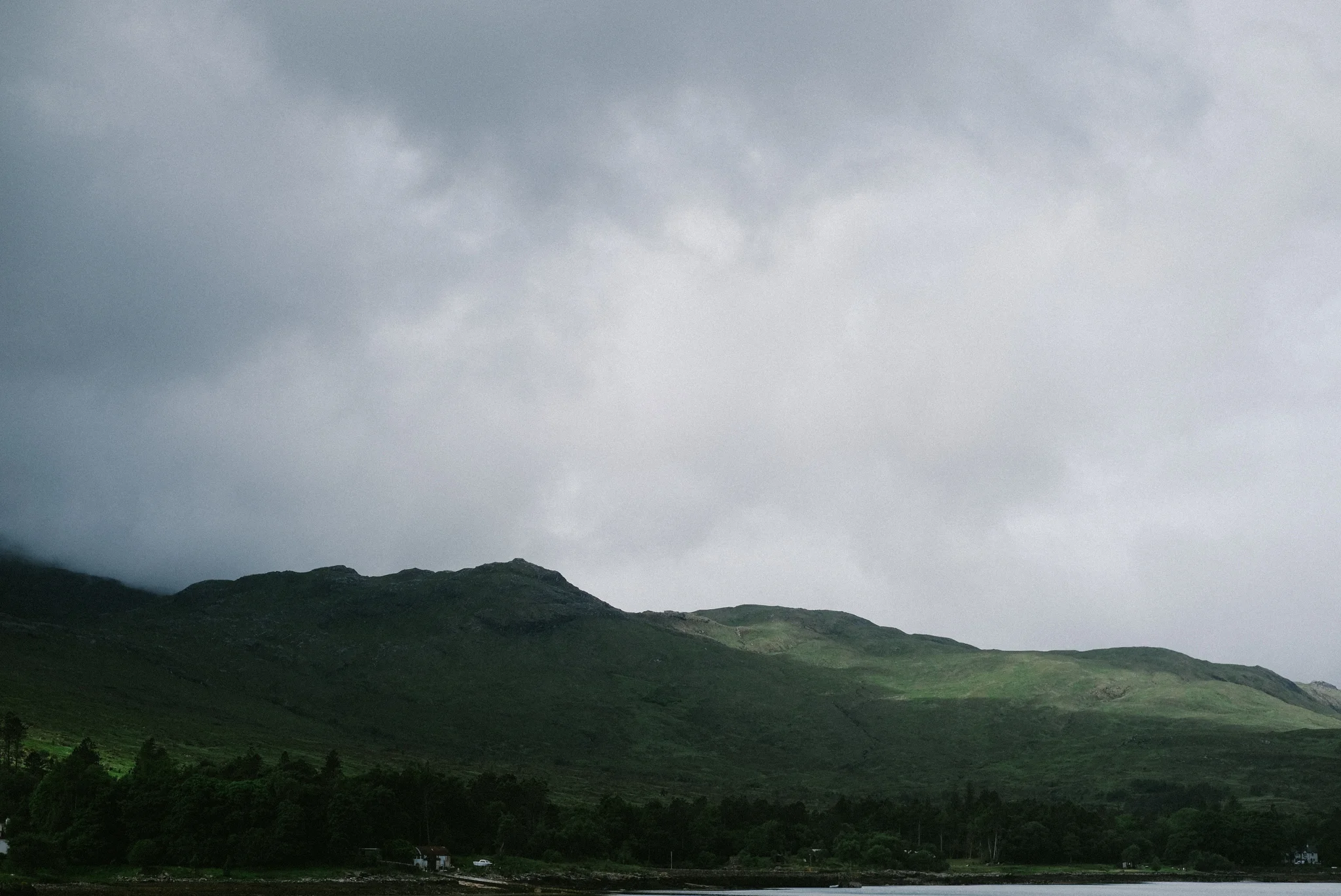 A landscape of green rolling hills with dark clouds overhead and a small house at the base of the hills.