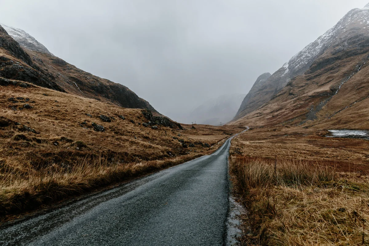 A winding paved road through a mountain valley with brown grassy hills and snow-capped peaks under a cloudy sky.