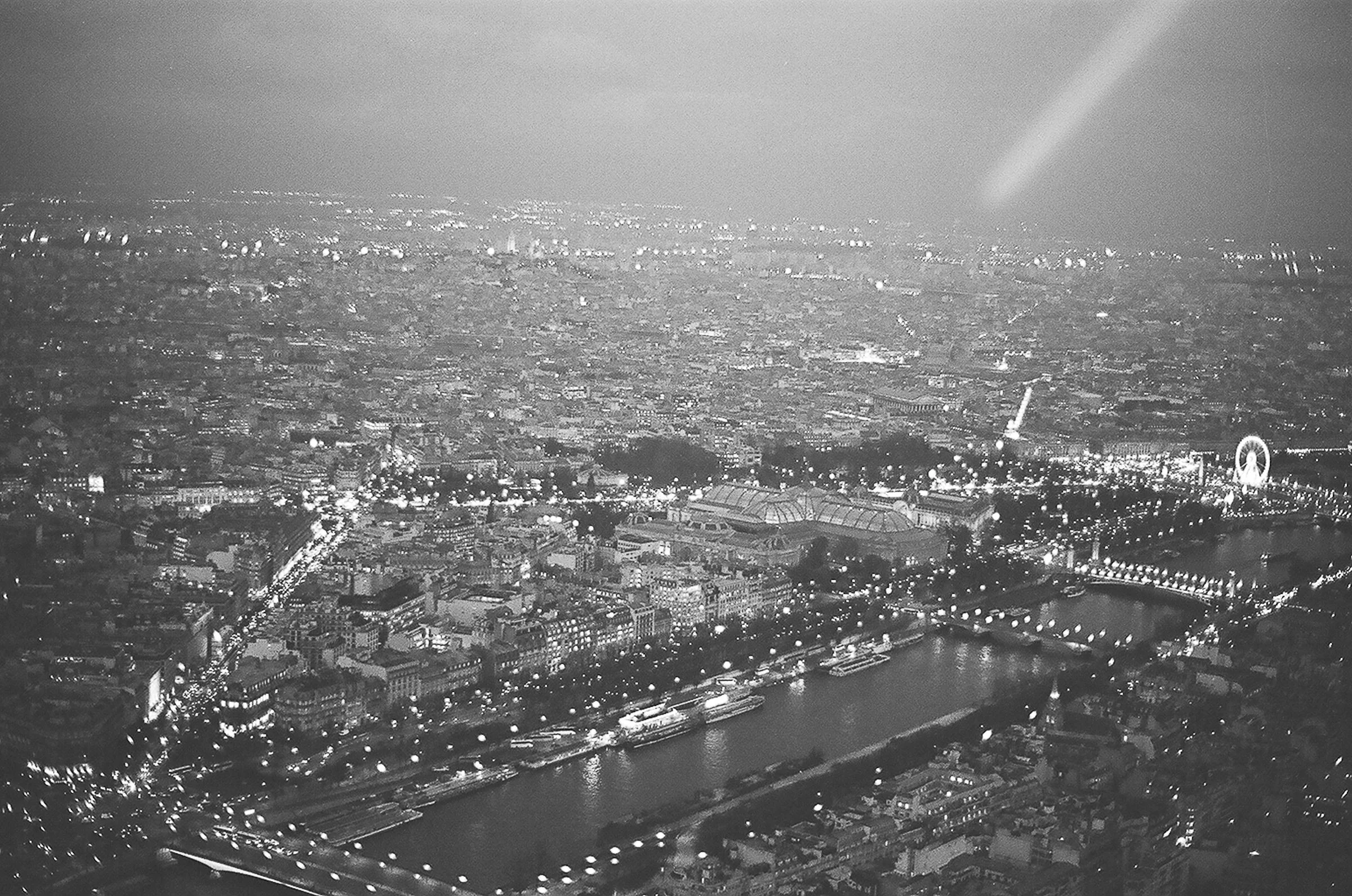 A black and white aerial night view of a city with illuminated buildings, streets, and a river running through it, viewed from a high vantage point.