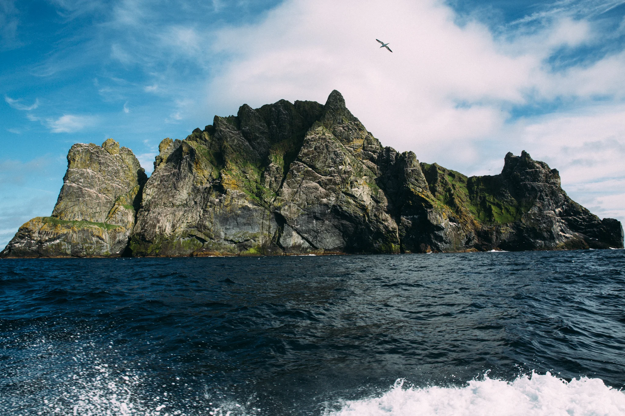 Large rugged rocky island with patches of greenery, seen from the water with waves in the foreground and a partly cloudy sky with bird in flight above.