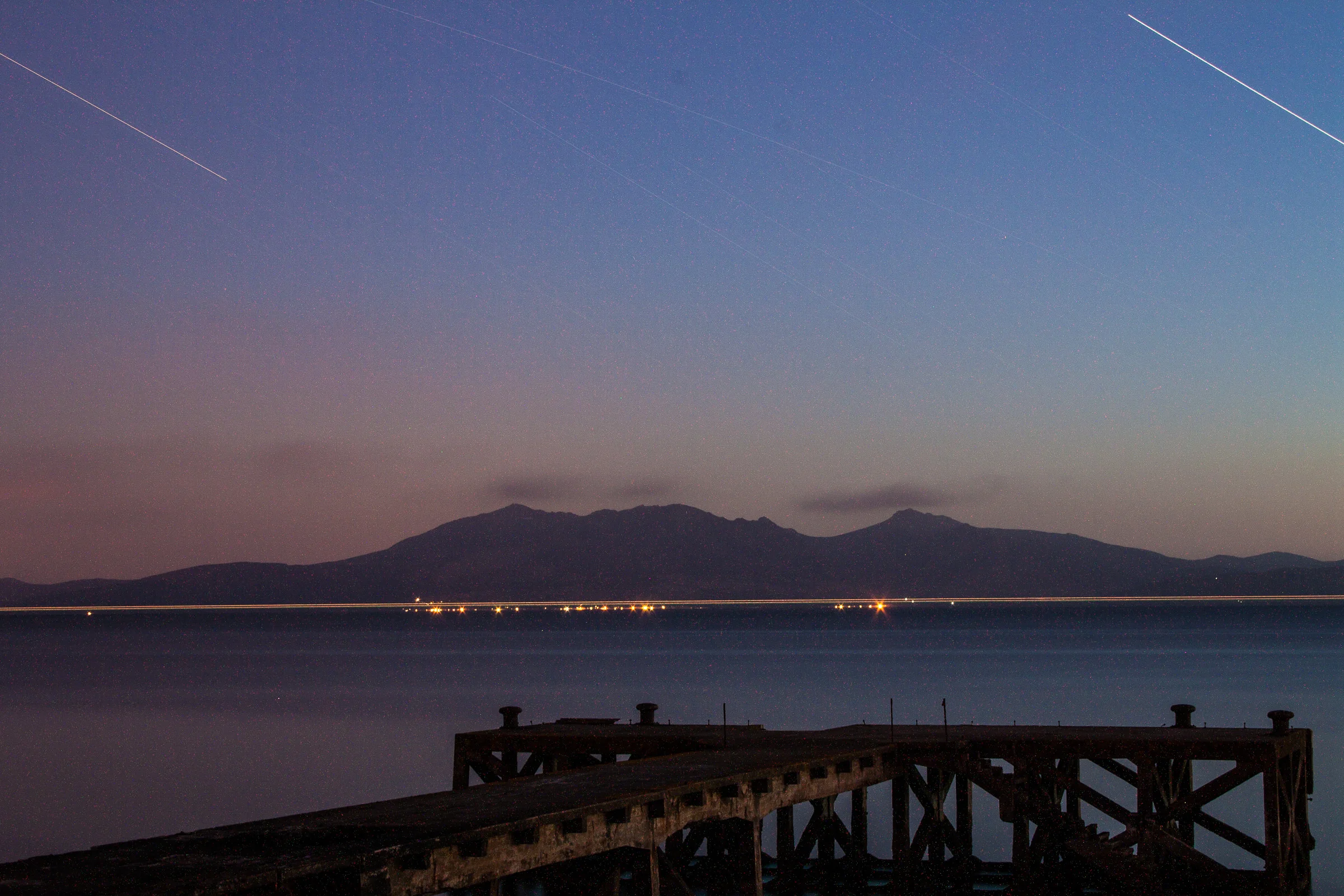 Nighttime view of a mountain range across a body of water with a wooden pier in the foreground and stars visible in the sky.