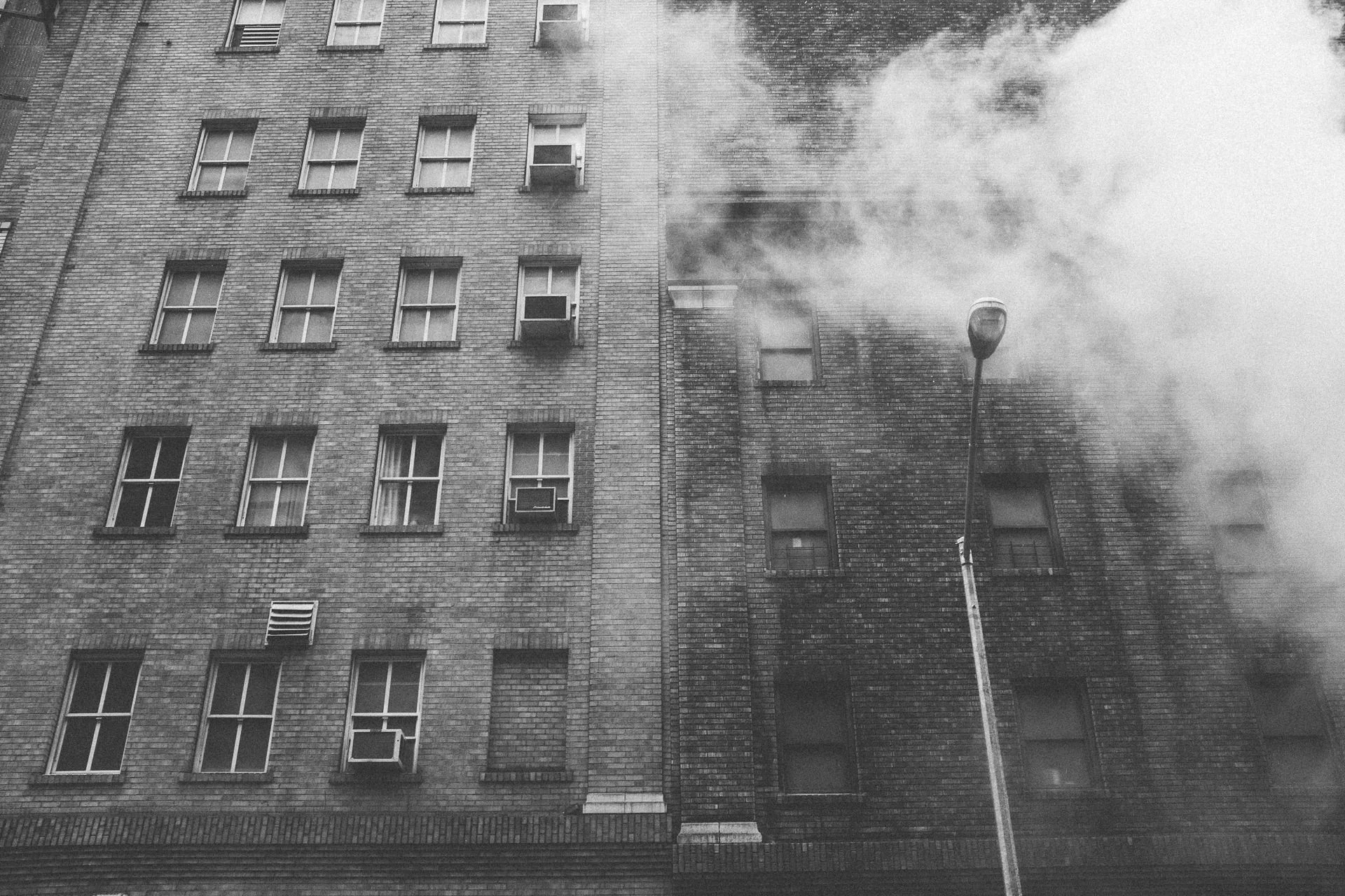 Black and white photo of a multi-story brick building with smoke billowing from a window, a streetlamp in front.