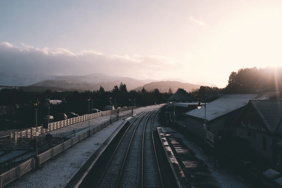A train station platform with train tracks, buildings, parked cars, and snow, surrounded by trees and mountains in the distance during sunset.