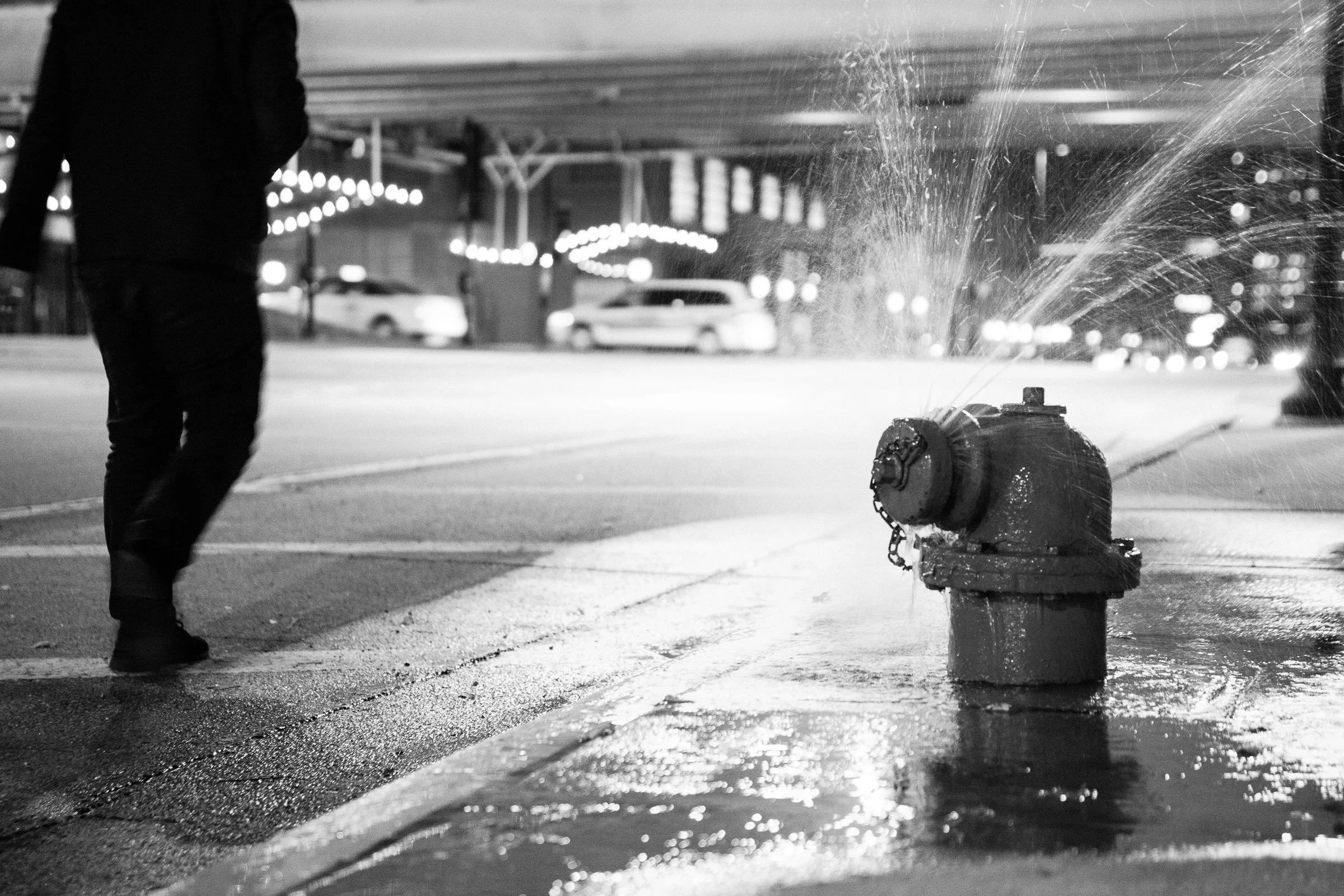 A person walking past a fire hydrant on a city street at night, with a fire hose spraying water onto the sidewalk, illuminated by streetlights and blurred cars in the background.