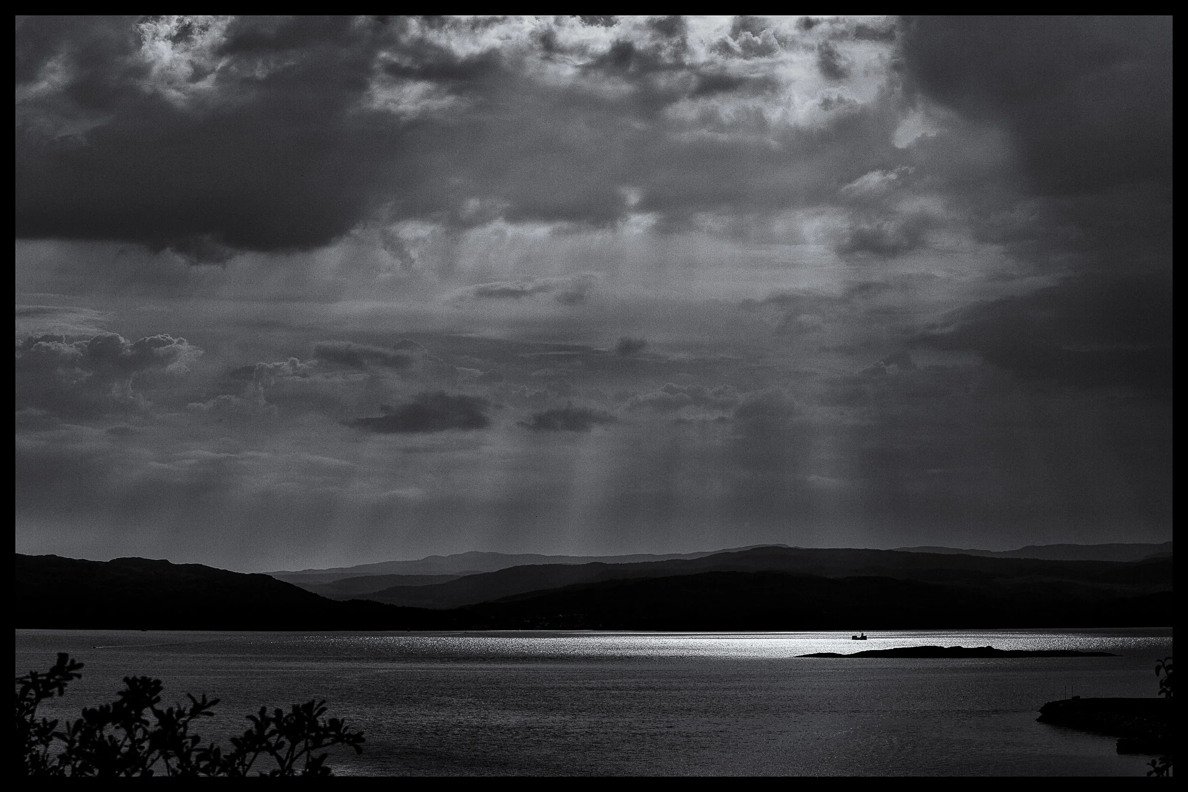 A black and white landscape photograph featuring a large body of water in the foreground, mountain ranges in the middle distance, and a dramatic cloudy sky above.