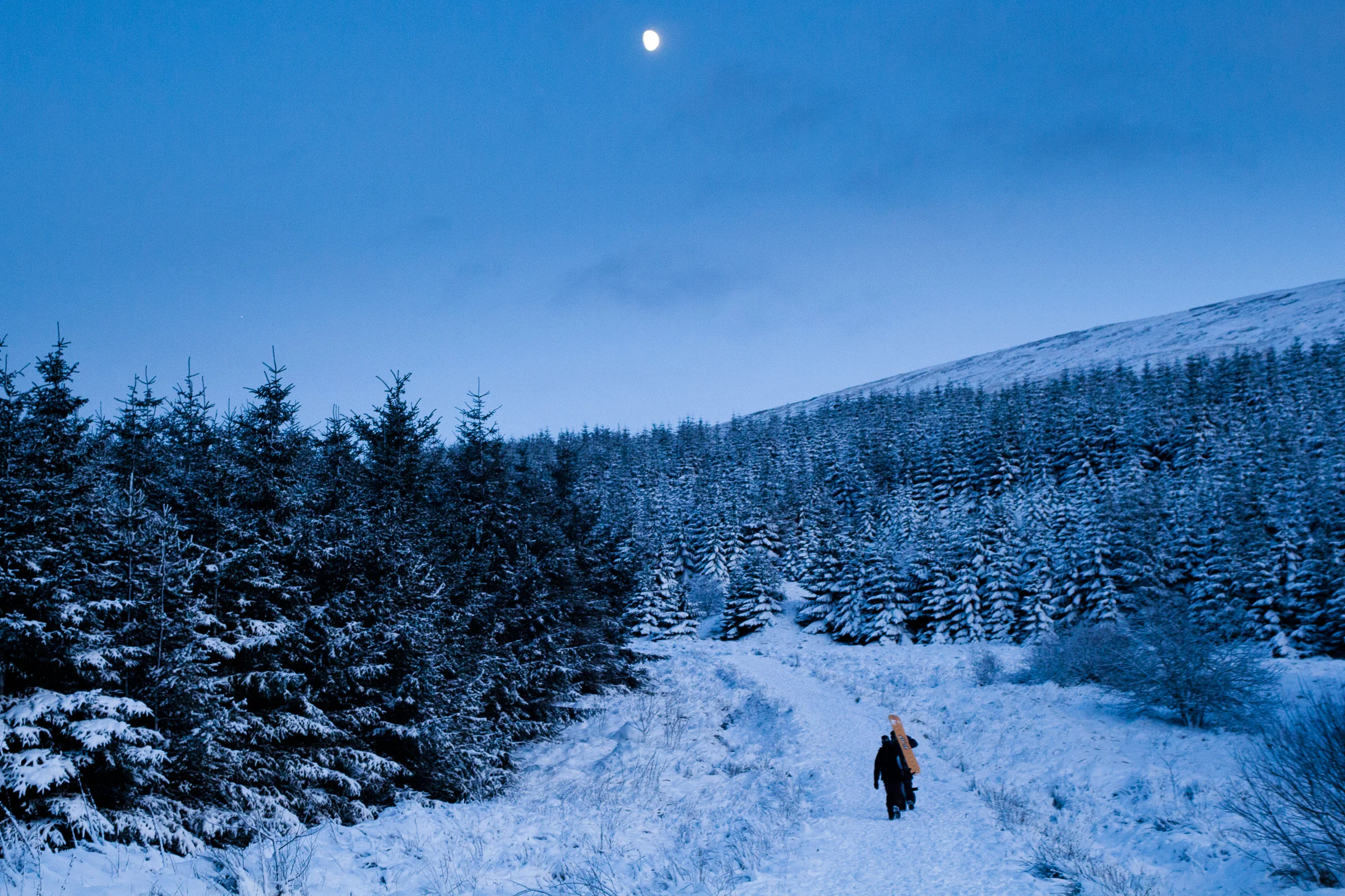 A snowy mountain landscape at dusk with a full moon in the sky, dense evergreen trees covered in snow, and a person walking with a snowboard on their back.