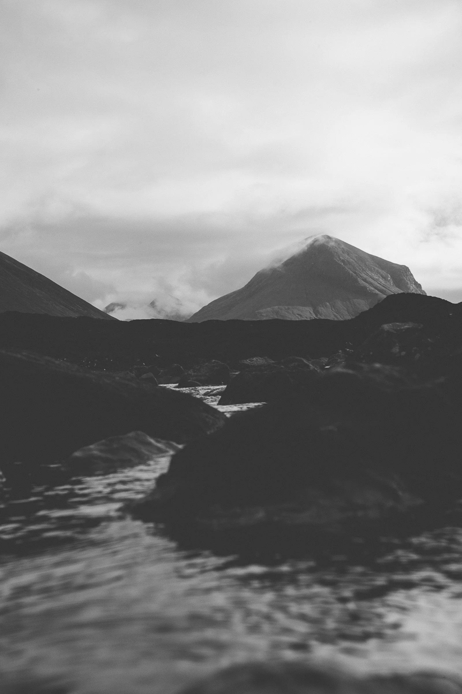 Black and white photo of a mountain with clouds around the peaks, viewed from a river with rocks in the foreground.