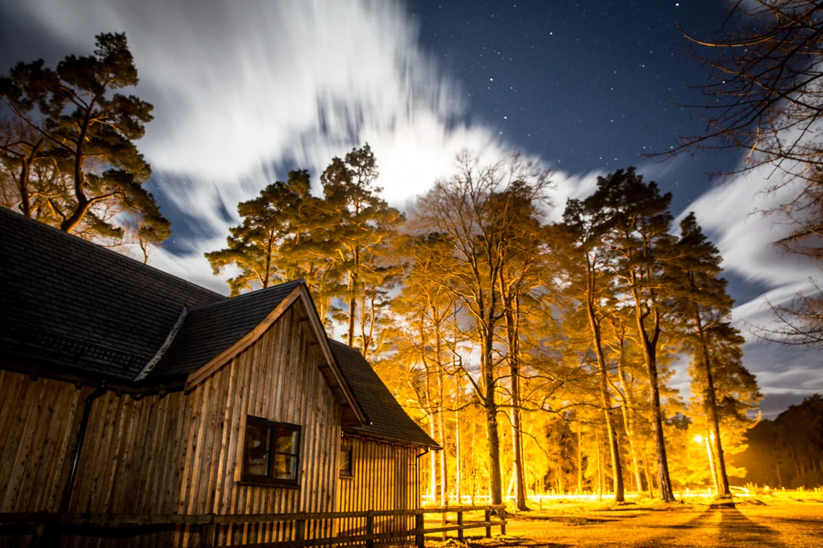 Nighttime scene of a rustic wooden house with a sloped roof surrounded by tall trees illuminated by warm yellow lights, with clouds moving across a starry sky.