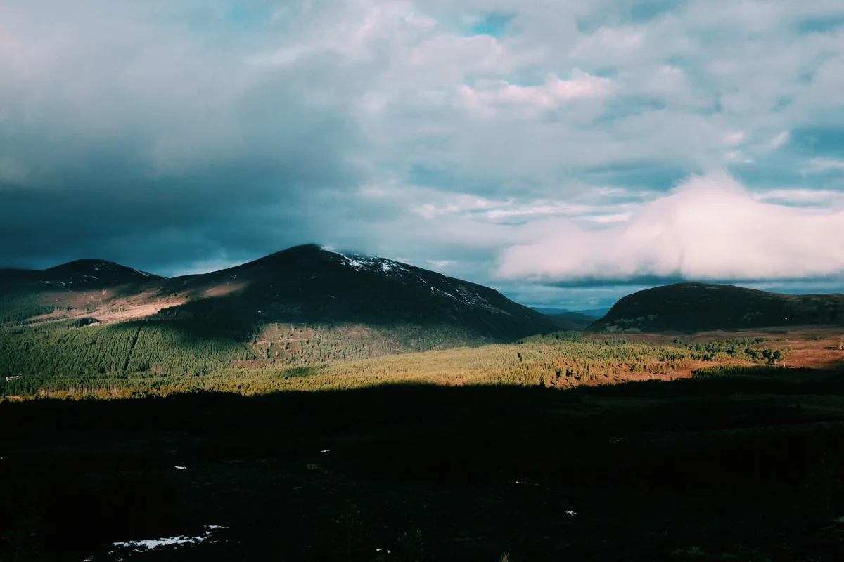 A landscape of rolling hills and mountains with patches of snow, green forests, and a partly cloudy sky.
