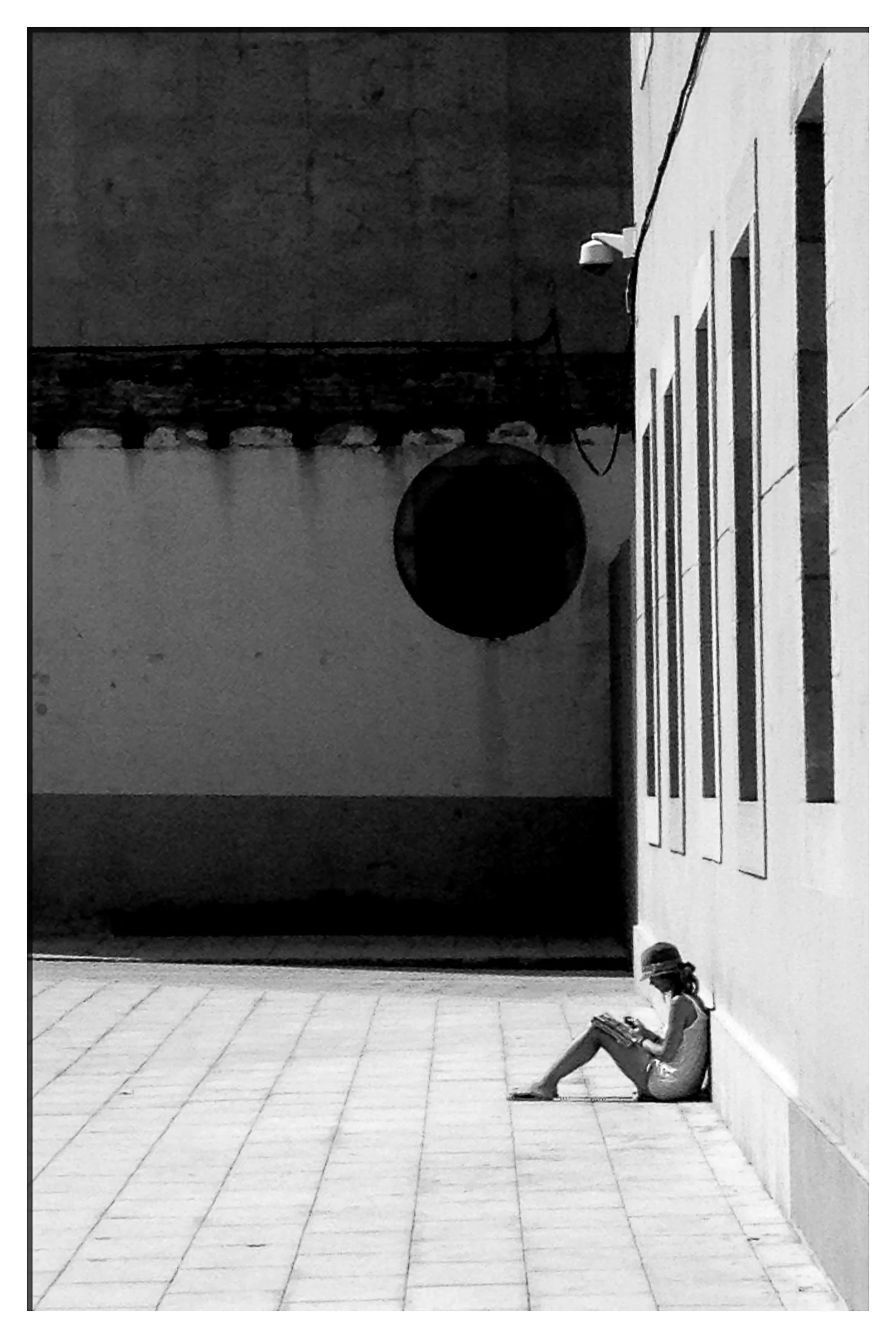 A girl sitting on the sidewalk reading a book near a white building with square windows, with a large black circle hanging on the wall and a girl in the background.