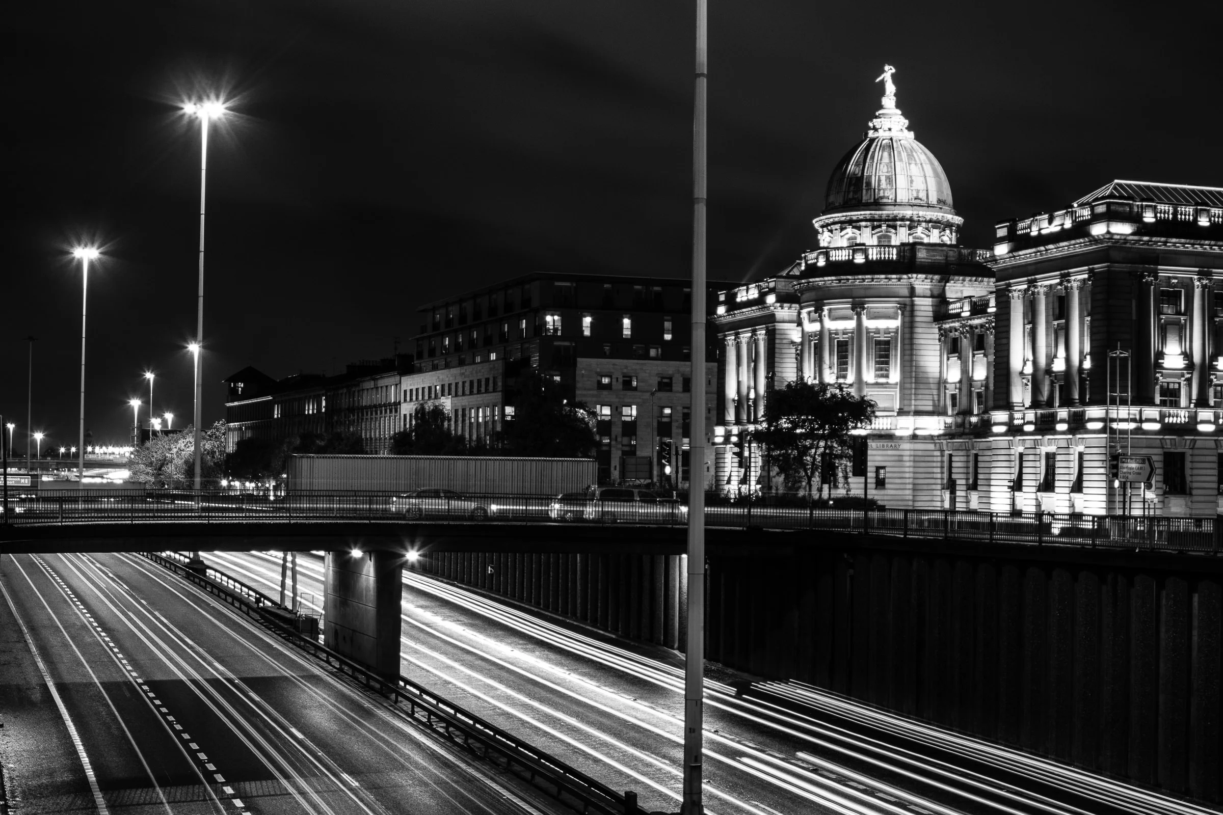Nighttime cityscape with illuminated historic building featuring a dome and statue on top, highway with light trails from moving vehicles, streetlights, and modern buildings in the background.