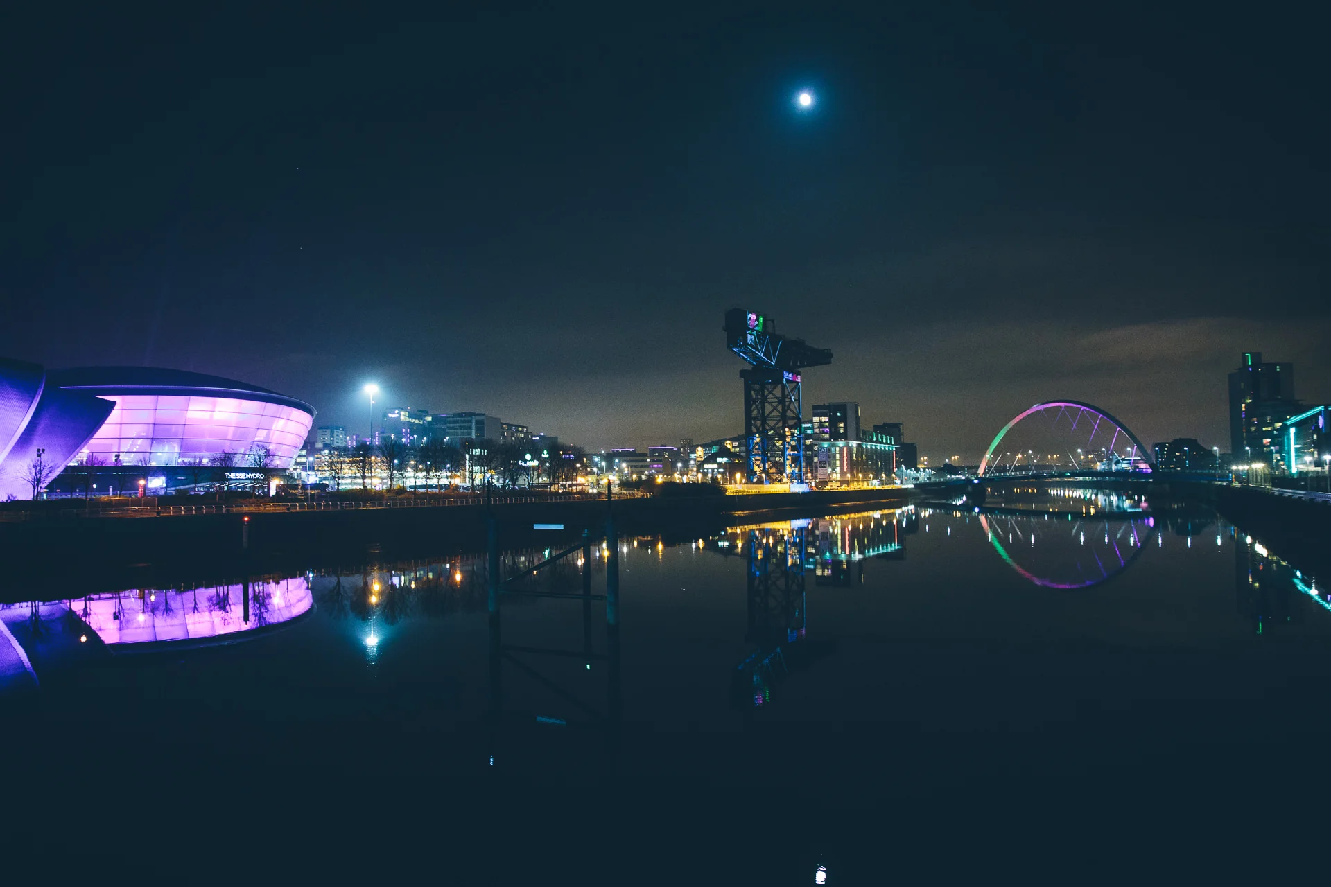 Nighttime cityscape featuring illuminated buildings, bridges, and a reflection on the water with a full moon in the sky.