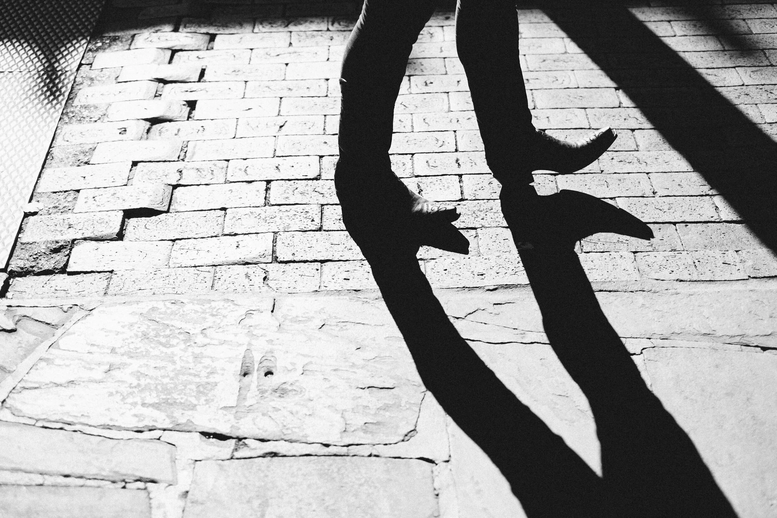 Black and white photograph of a person walking on a brick sidewalk, casting a long shadow due to bright sunlight.
