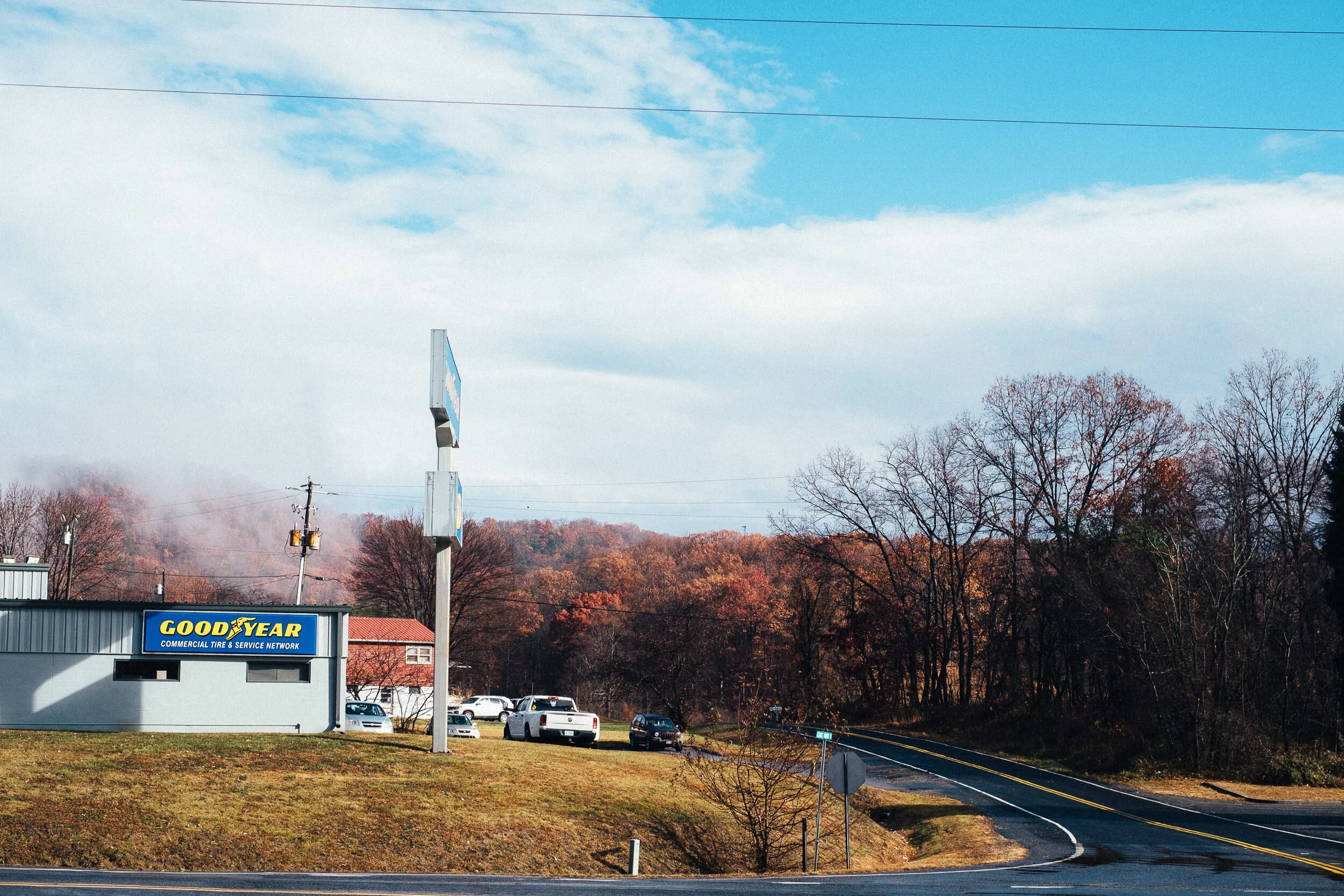 A commercial area with a sign for Goodyear, cars parked nearby, a tall light pole, and a scenic background of trees with autumn foliage under a partly cloudy sky.