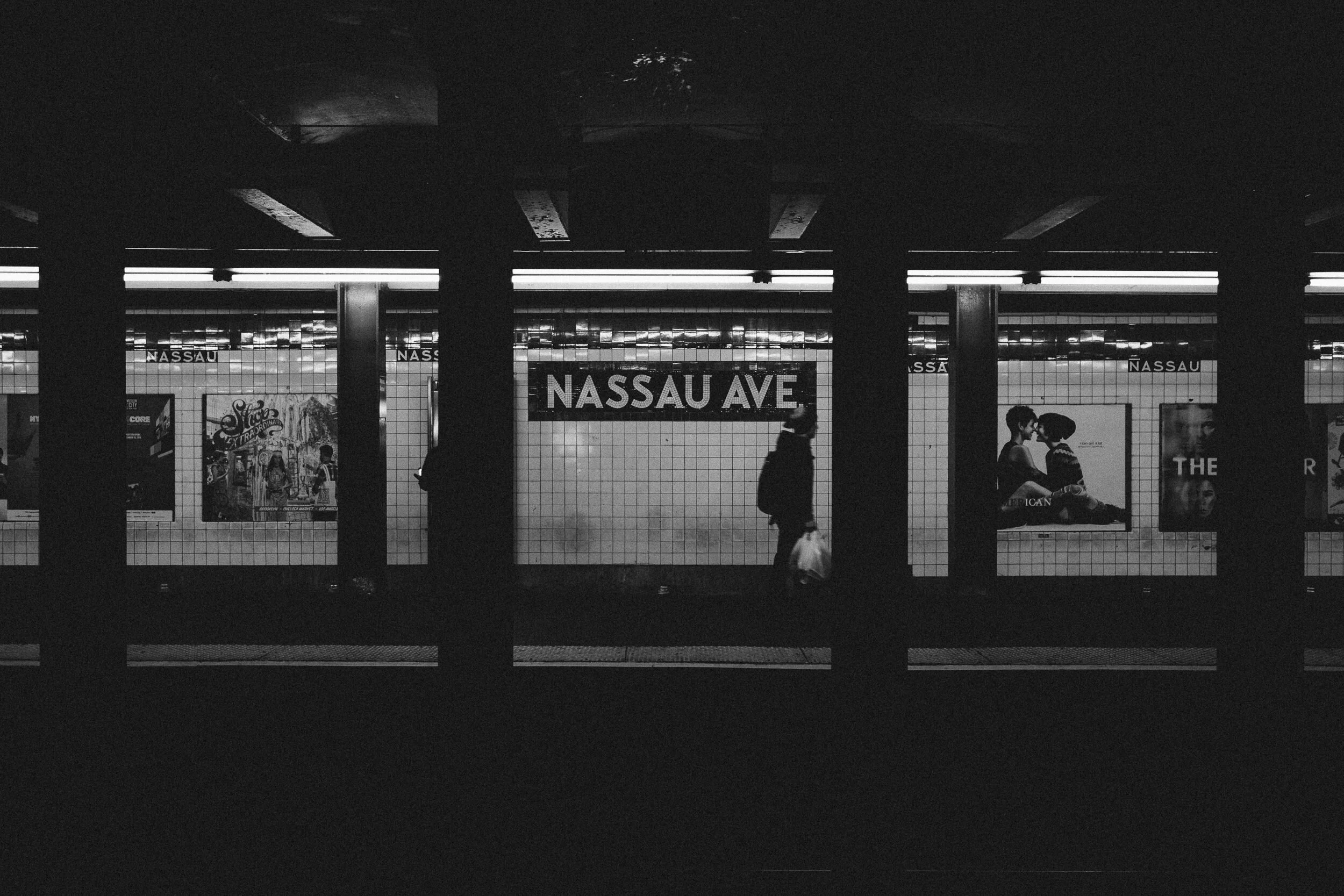 Underground subway station with a sign reading 'Nassau Ave' and several people walking past tiled walls with advertisements.