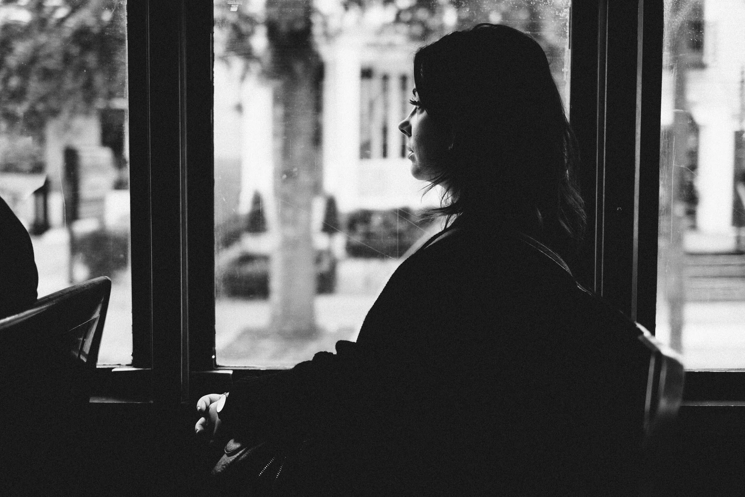 A black and white photo of a woman sitting in a waiting area, looking out a window at an urban street scene.