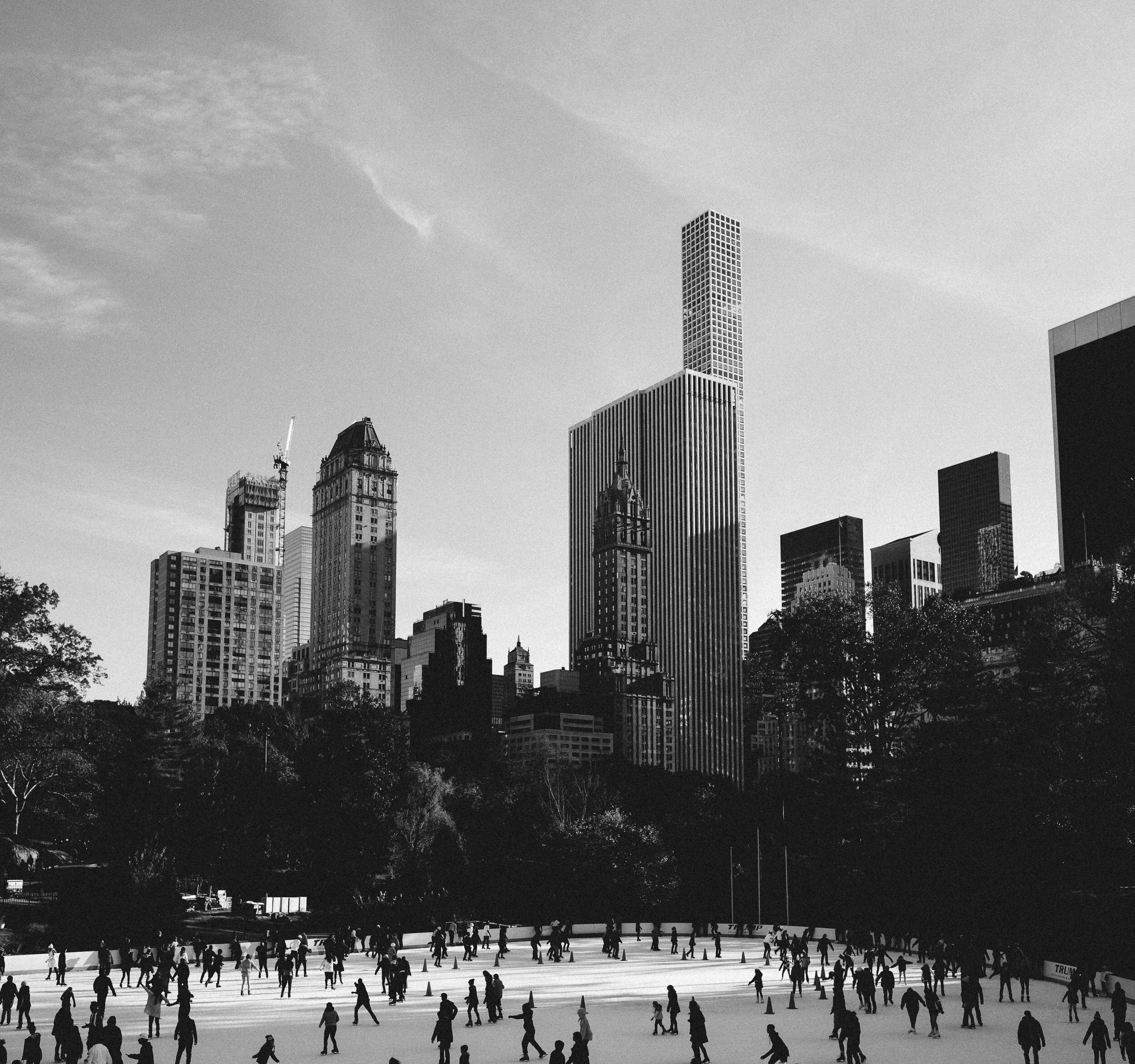 Black and white photograph of an outdoor ice skating rink filled with many skaters, with tall city buildings in the background and trees in the foreground.