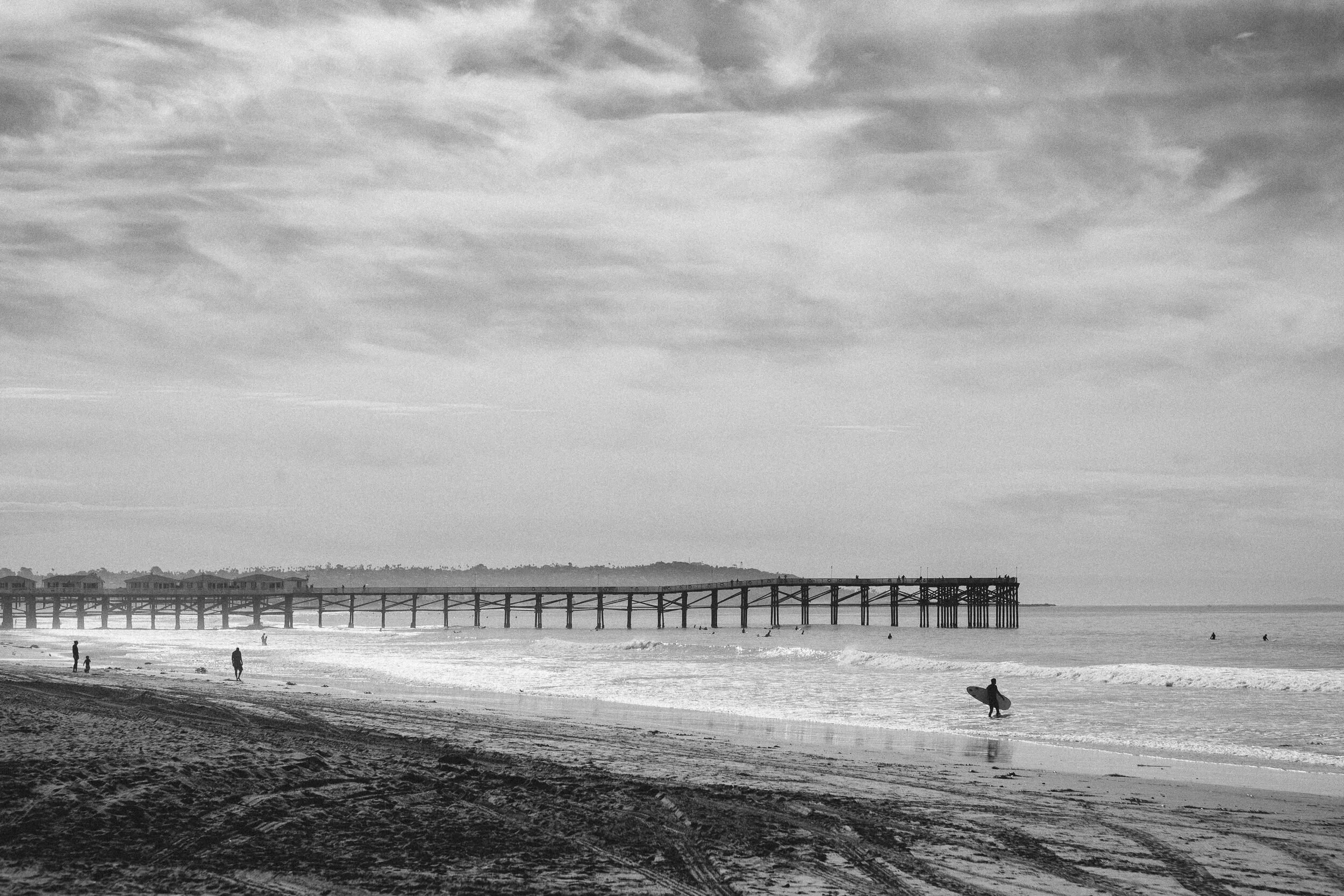 A black-and-white photo of a beach with a pier extending into the water. People are walking and surfing on the beach, and the sky is cloudy.