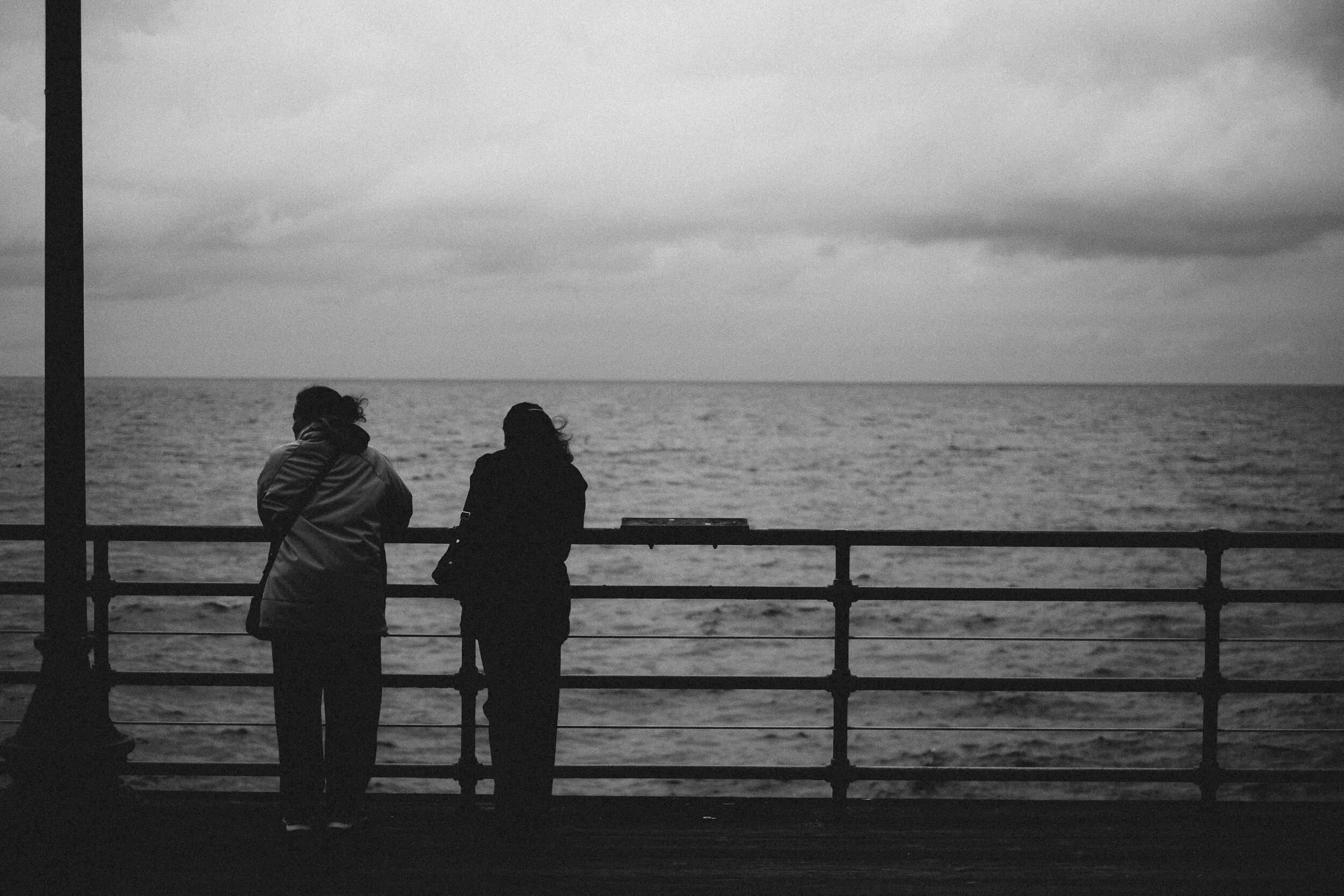 Two people sitting on a bench by the water under a cloudy sky.