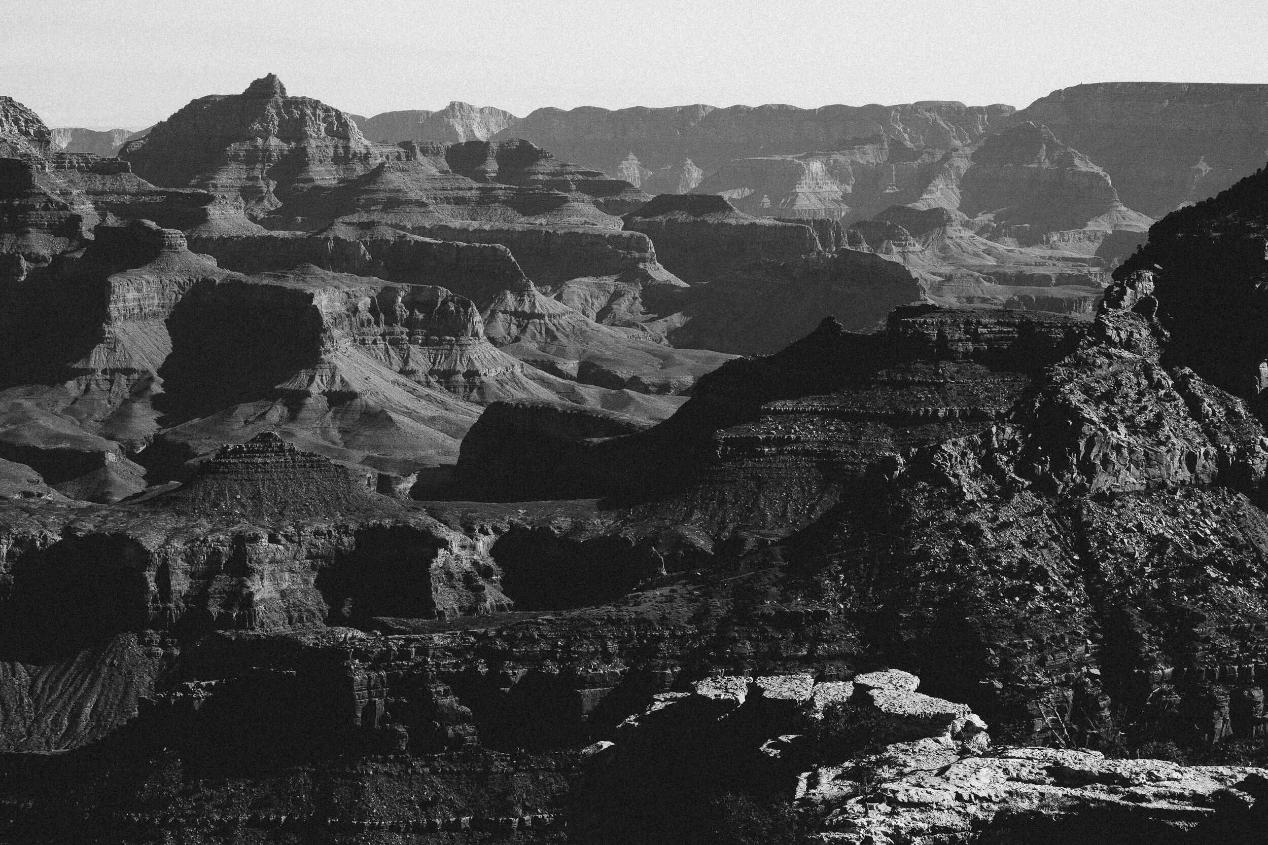 Black and white photograph of the Grand Canyon with layered rock formations and rugged cliffs.
