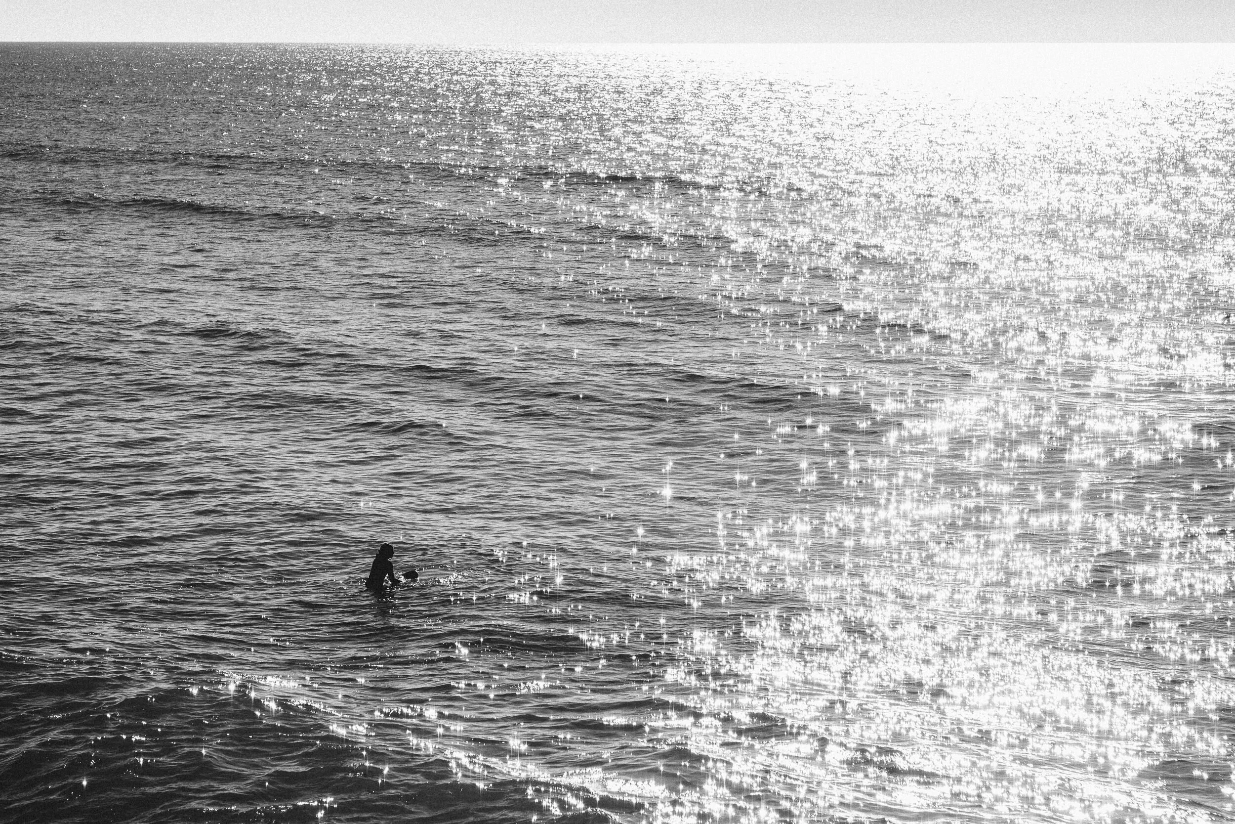 Silhouette of person holding a ball while standing in the ocean at sunset, with sparkling water and horizon in the background.