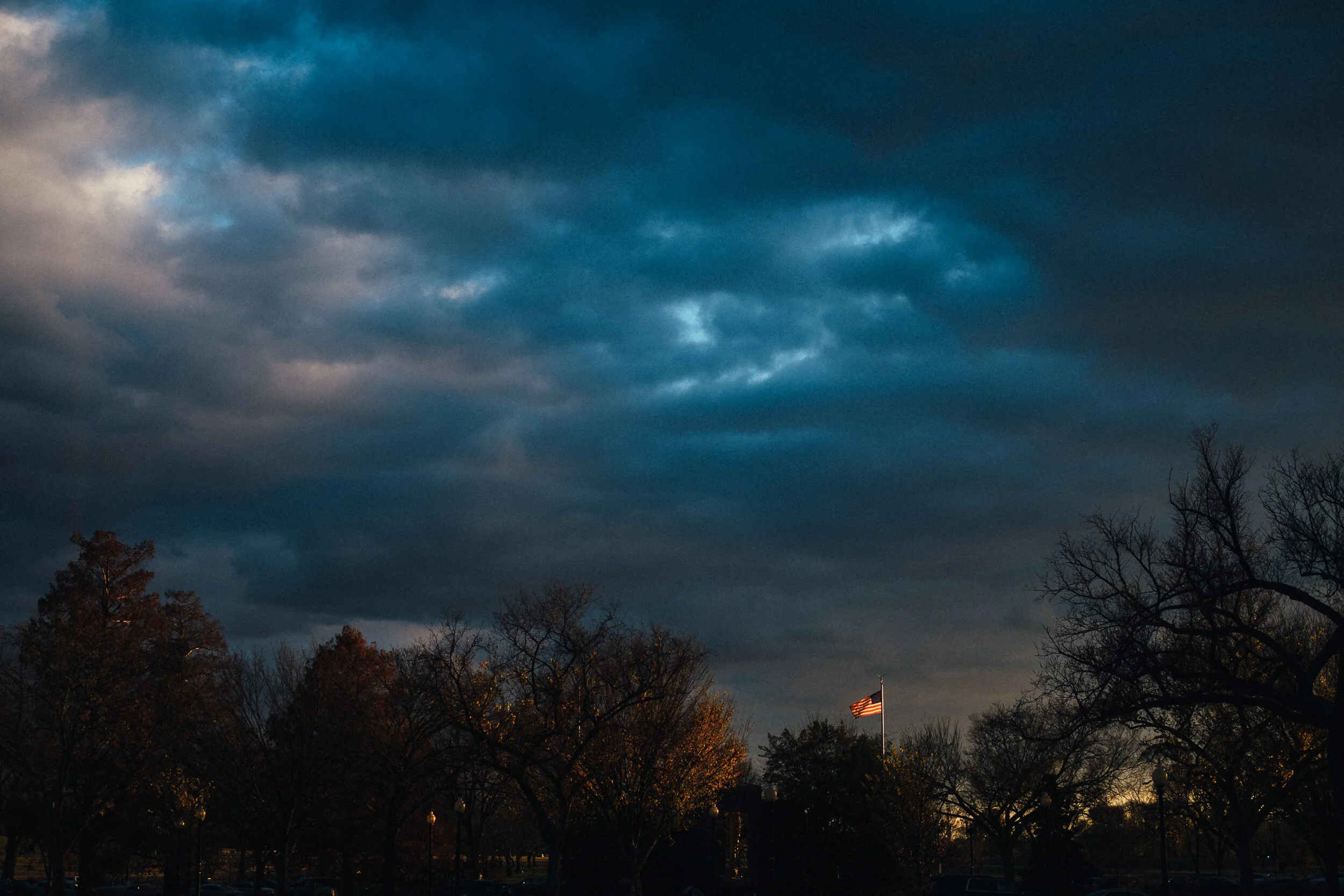 Dark cloudy sky over trees and an American flag in the distance.