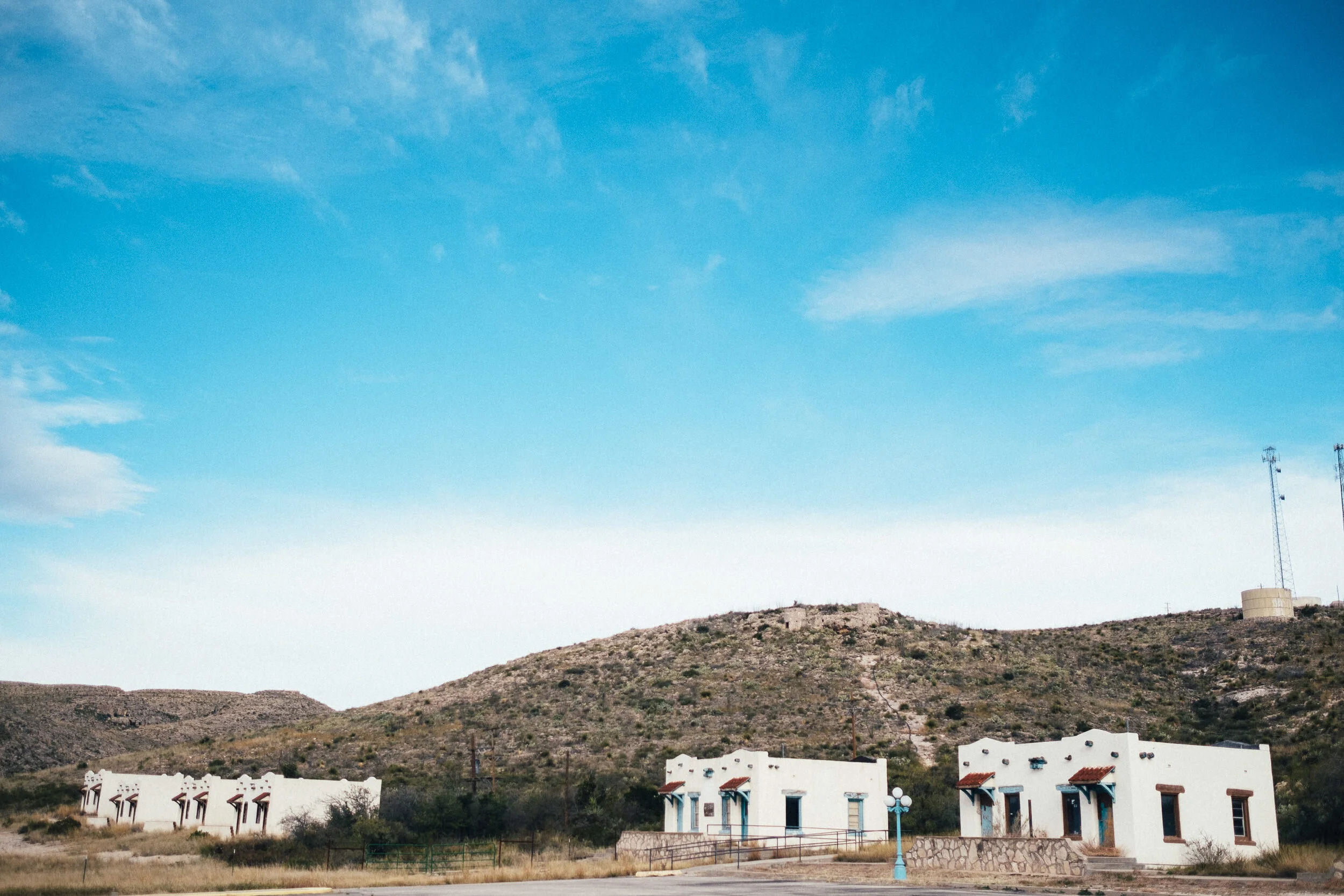 A landscape view of a desert area with white buildings in the foreground, a hill with sparse vegetation, and a mountain in the background under a blue sky with some clouds.