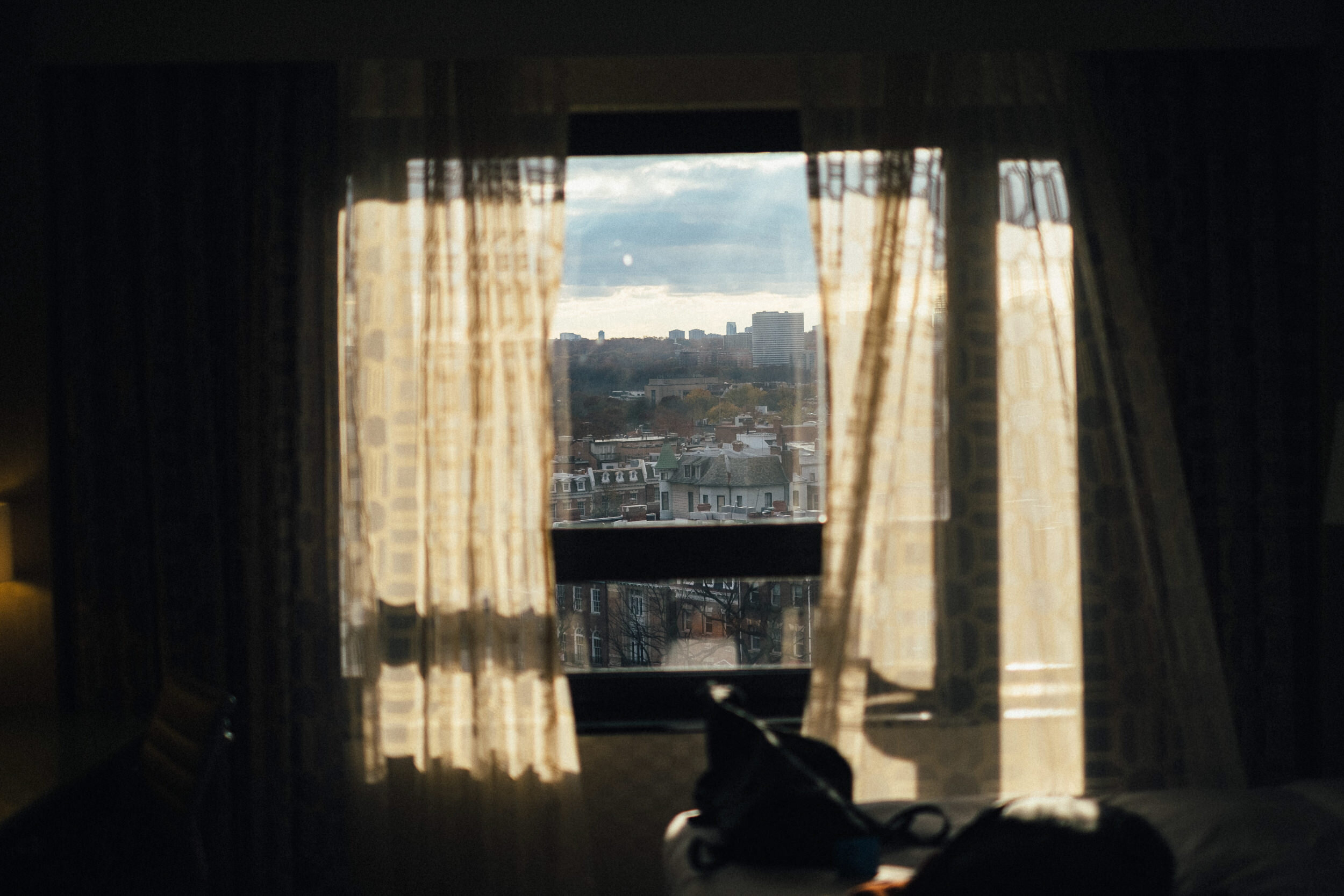 View through a window with beige curtains, showing a cityscape with old and new buildings under a cloudy sky.