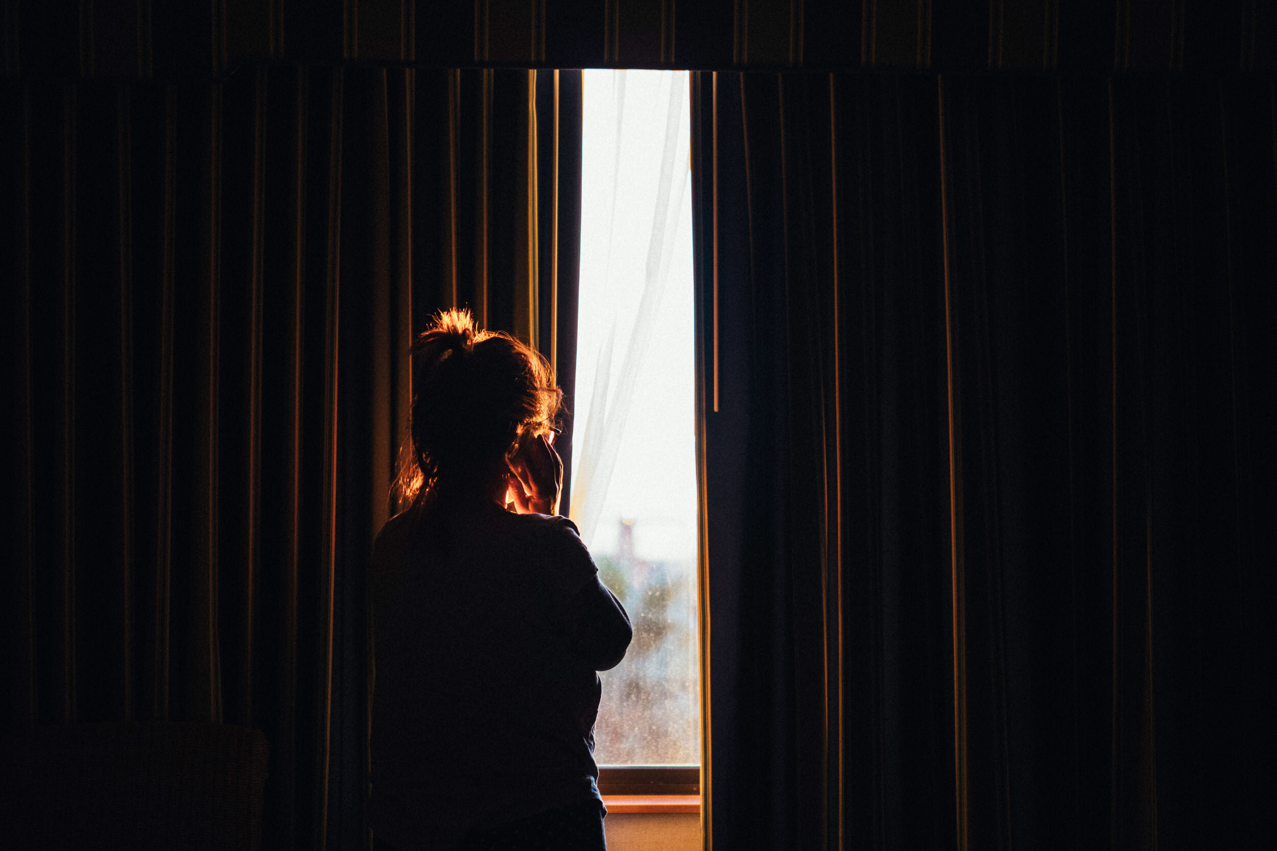A person with a ponytail looking out of a window with dark curtains, sunlight illuminating the person's hair and part of the face, with a distant cityscape or horizon visible outside.