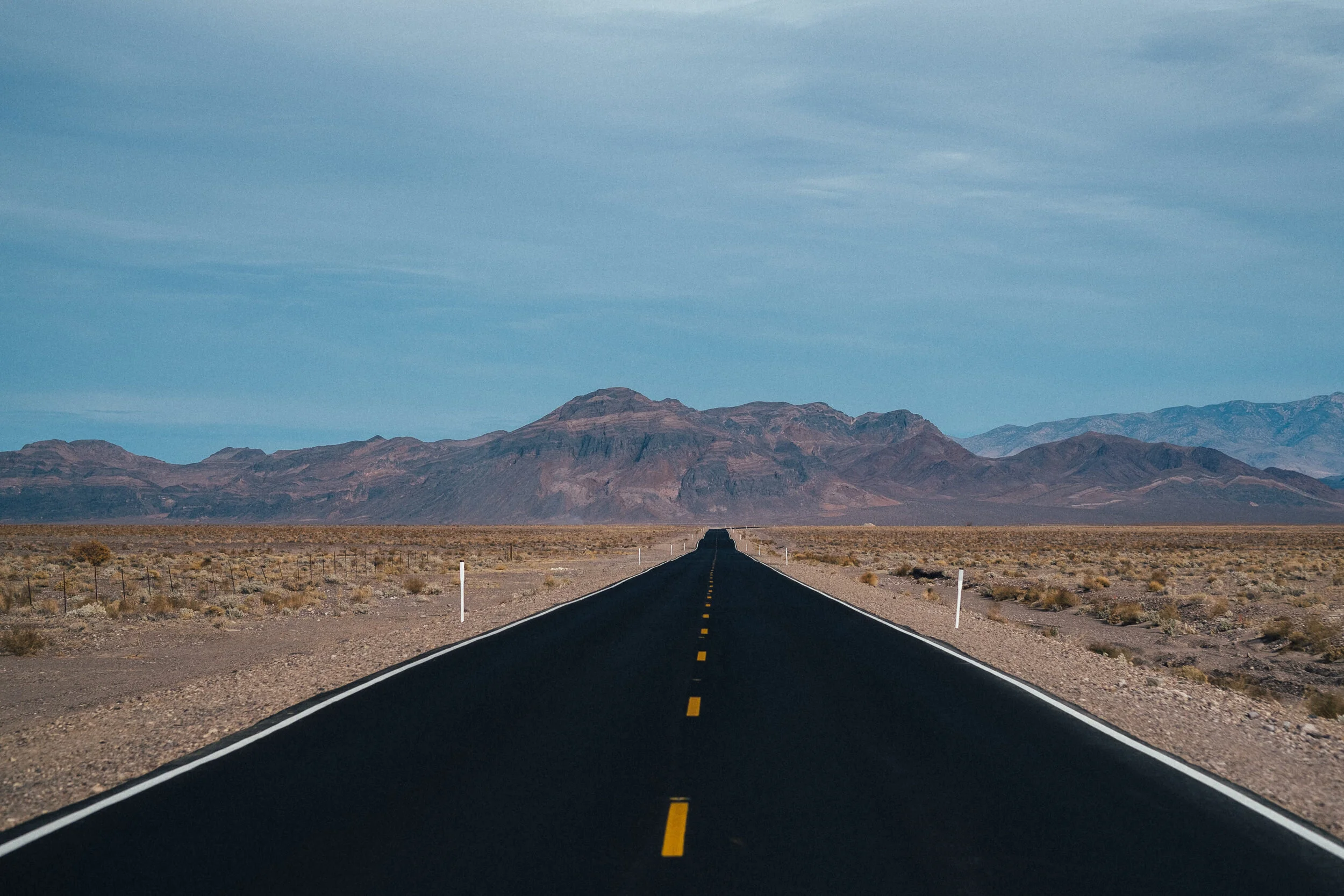 A long empty road stretching toward distant mountains under a partly cloudy sky in a desert landscape.