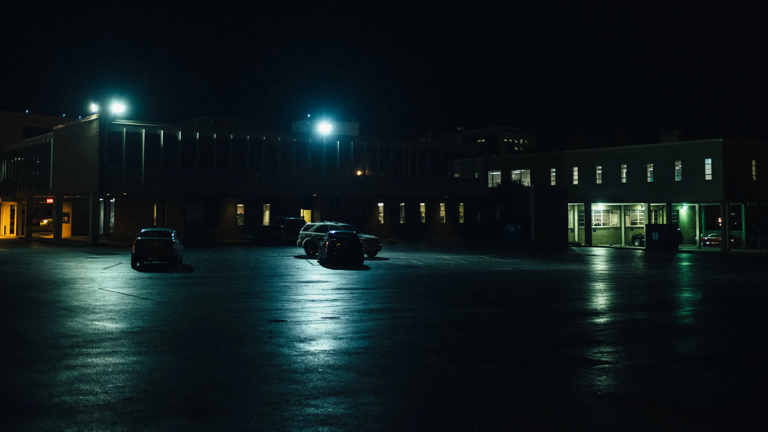 Nighttime view of a mostly empty parking lot outside a building, with a few cars parked and illuminated windows, under dark sky.
