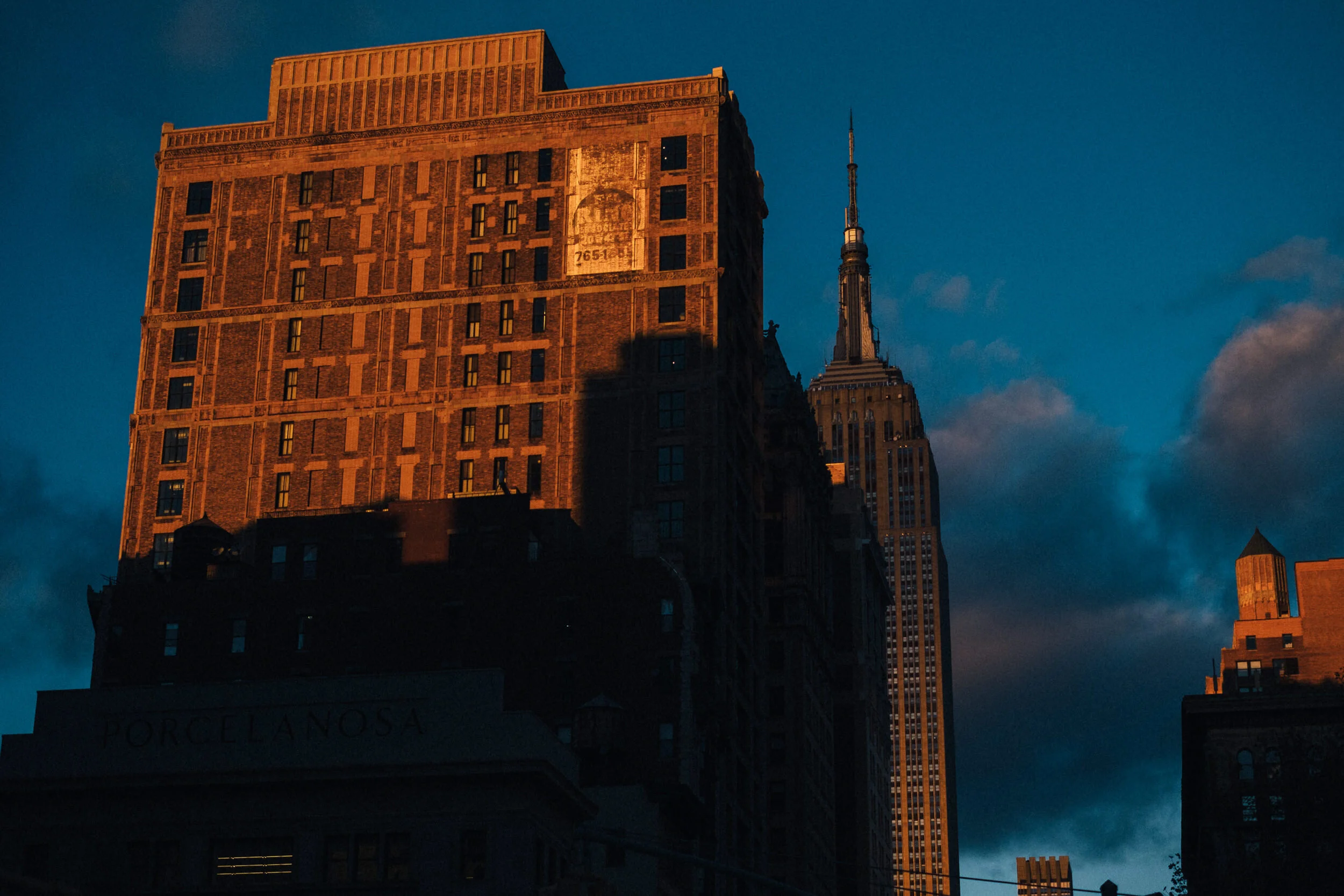 A cityscape with a tall brown brick building in the foreground, illuminated by warm sunlight, with a visible billboard and several windows. In the background, the Empire State Building is visible against a darkening evening sky.
