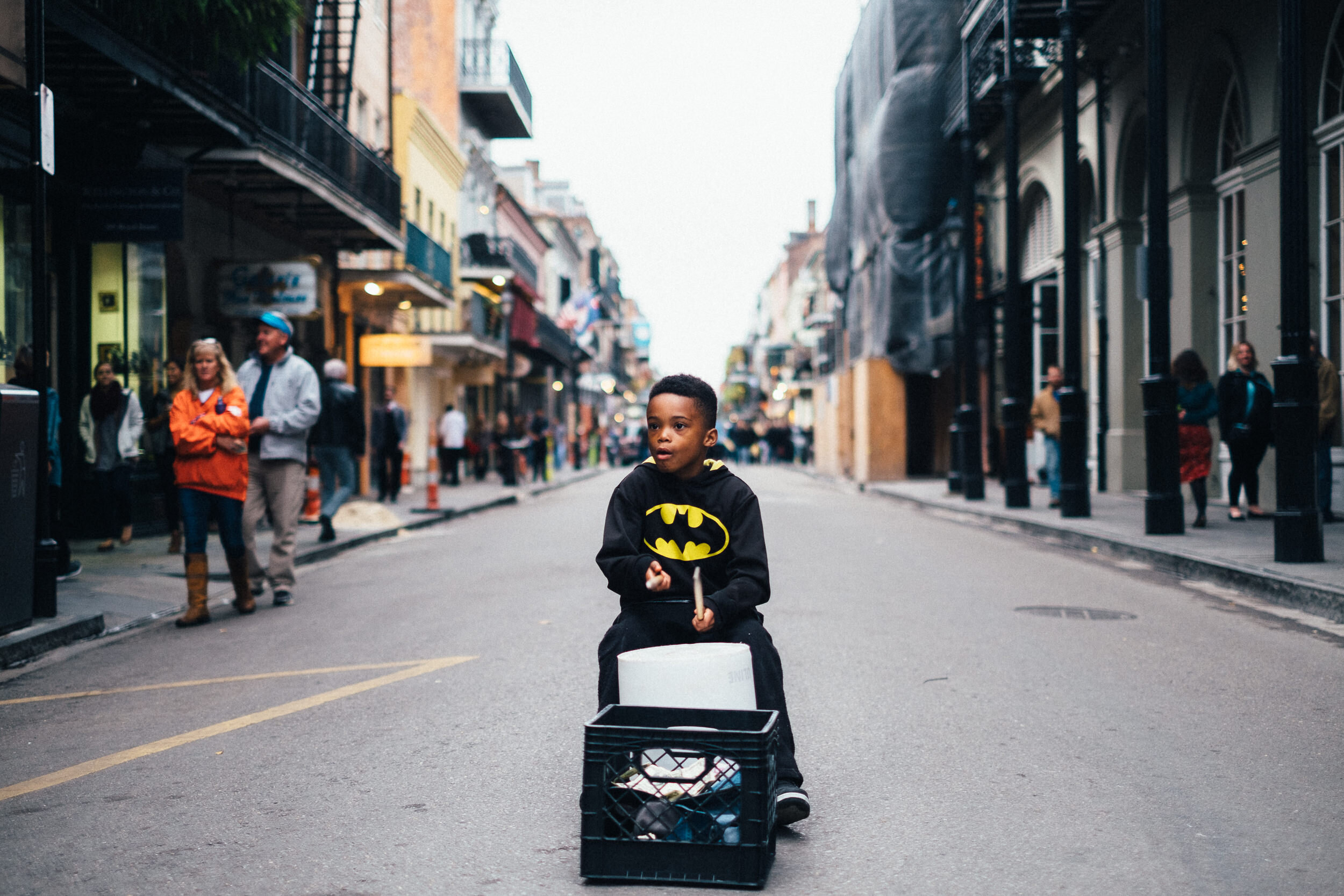 A young boy dressed in a Batman shirt rides a scooter in the middle of a city street, with pedestrians on either side, and buildings lining the street during daytime.