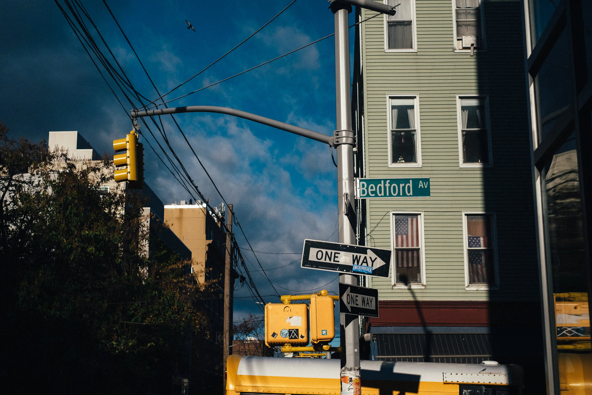 Street scene showing traffic lights, street signs for Bedford Avenue, and one-way signs at an intersection next to a green multi-story building with yellow and American flags in the windows. A bird is flying in the sky.