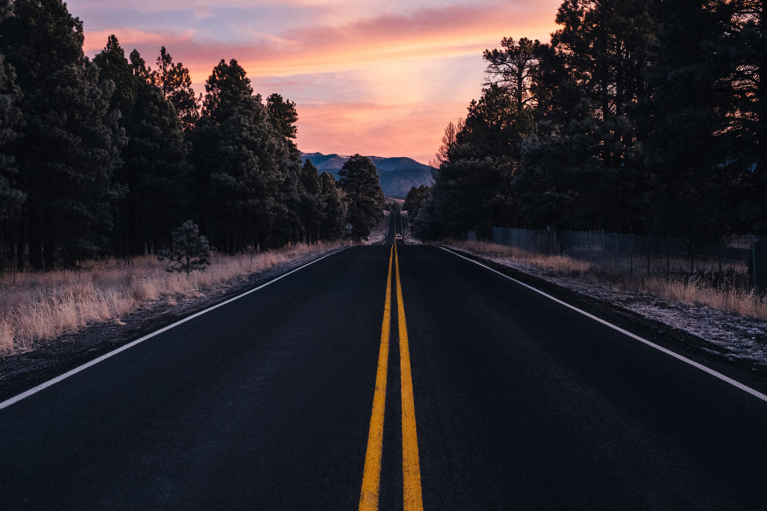 A long asphalt road with yellow double lines running through a forested landscape at sunset, with mountains visible in the background and a colorful sky.