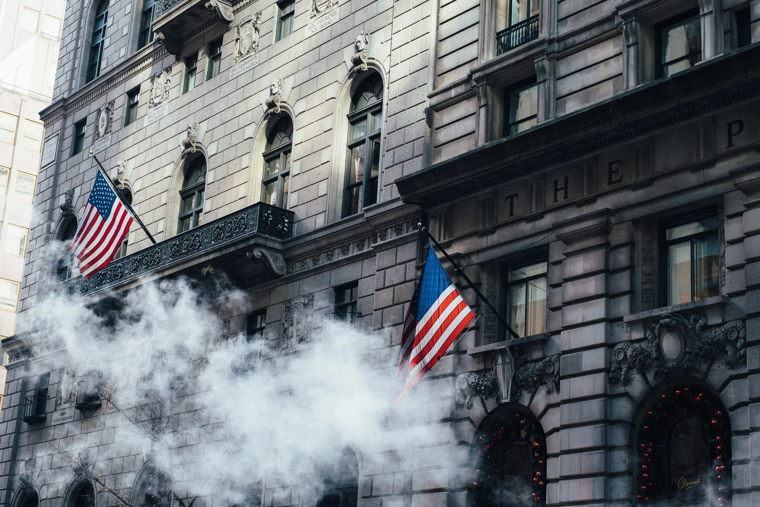 The facade of a tall, historic building decorated with ornate architectural details, with American flags hanging from its balconies. Smoke is billowing from the lower part of the building.