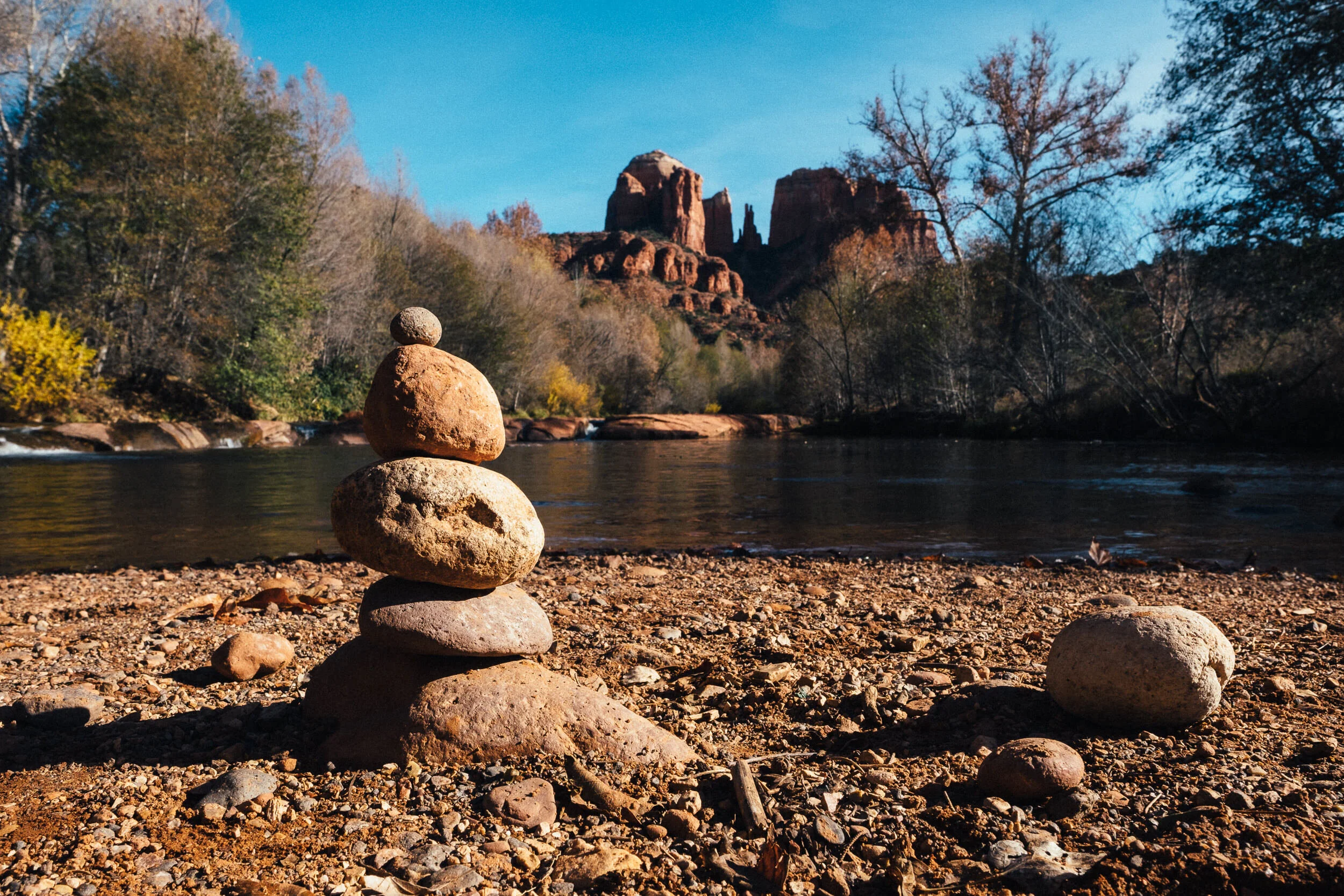 Stacked stones on a rocky riverbank with trees and red rock formations in the background under a blue sky.
