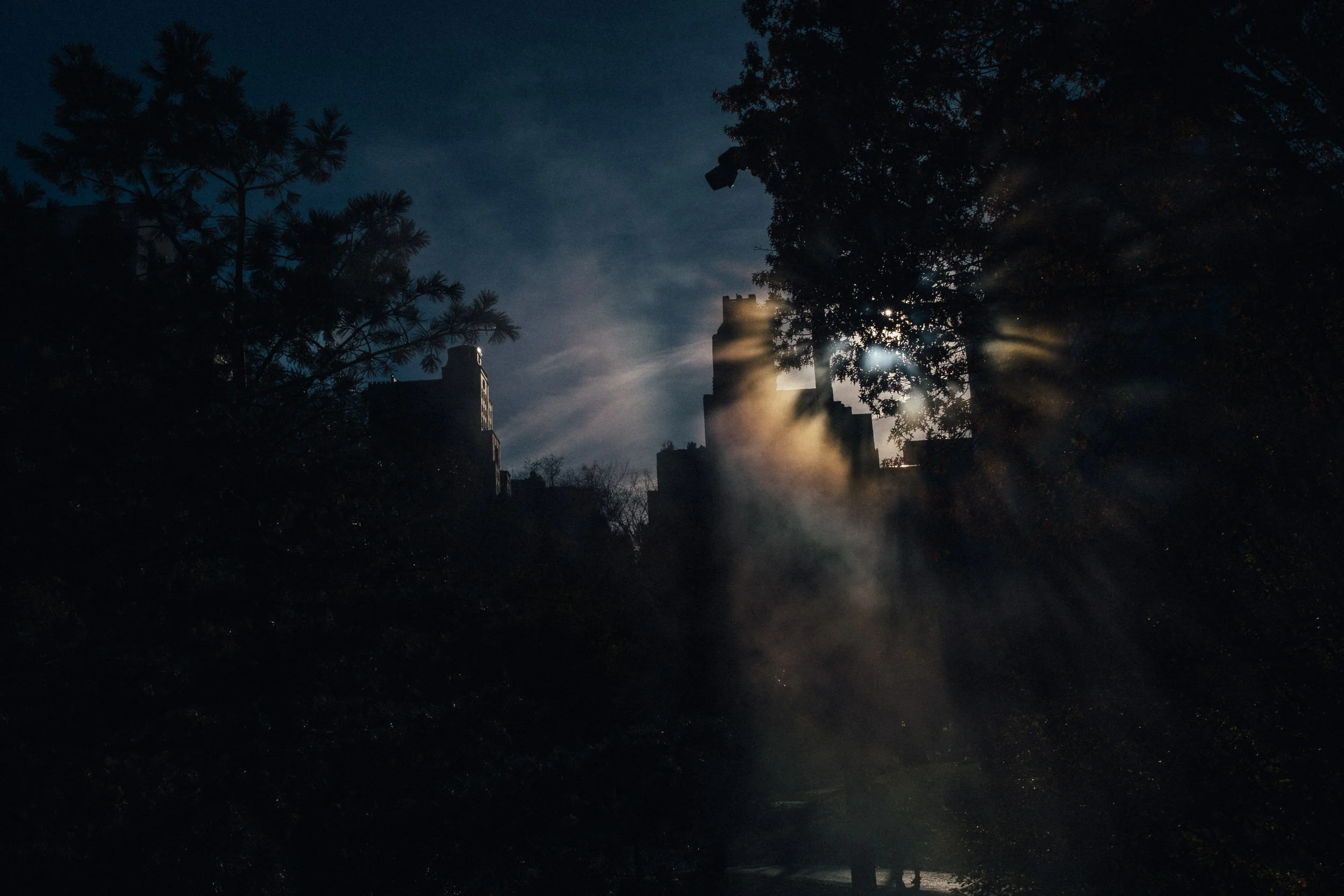 Nighttime scene with dark trees and smoke, with sunlight shining through, illuminating building rooftops in the background.