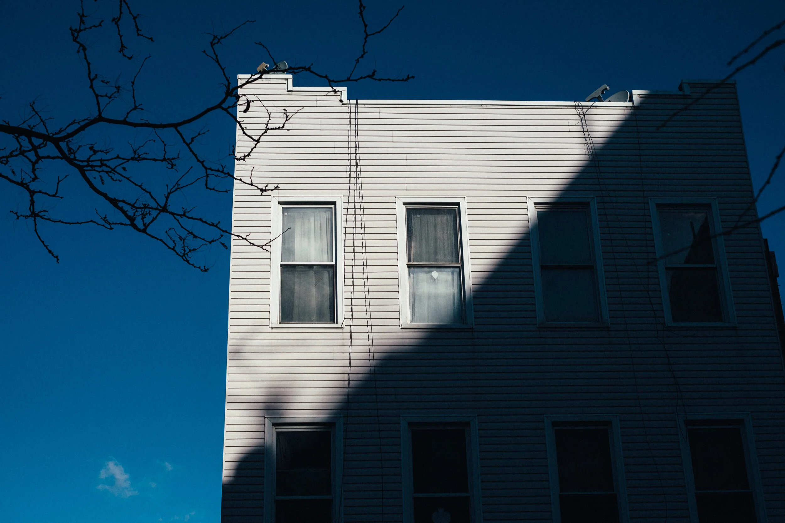 A white multi-story house with horizontal siding, six windows, and a shadow on the right side, with leafless tree branches in the foreground and a clear blue sky in the background.