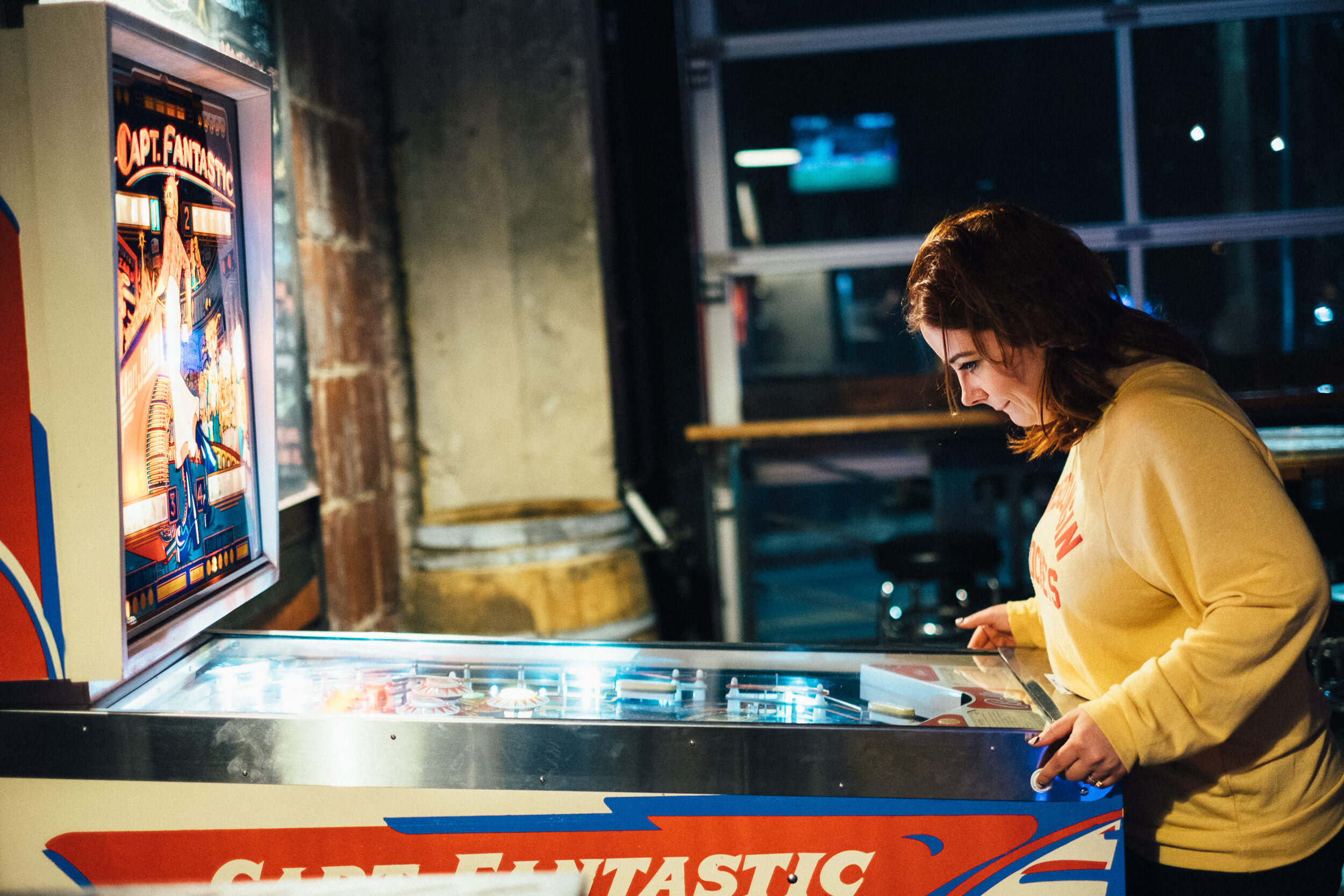 A young woman in a yellow sweatshirt playing pinball at a game arcade, with the pinball machine titled 'Captain Fantastic' visible on the side.