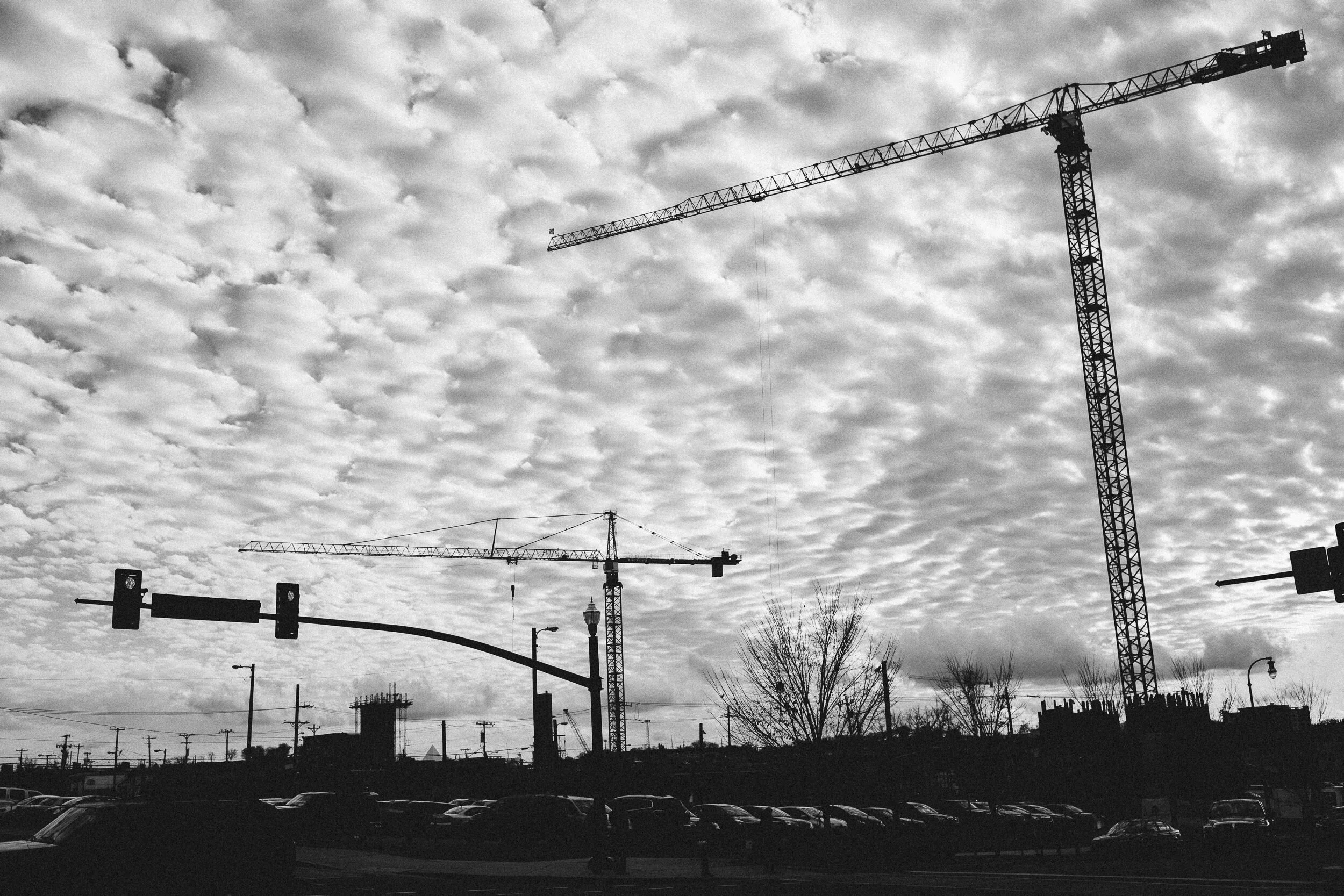 Black and white photo of a cityscape featuring multiple construction cranes, streetlights, power lines, cars in a parking lot, and an overcast sky with textured clouds.