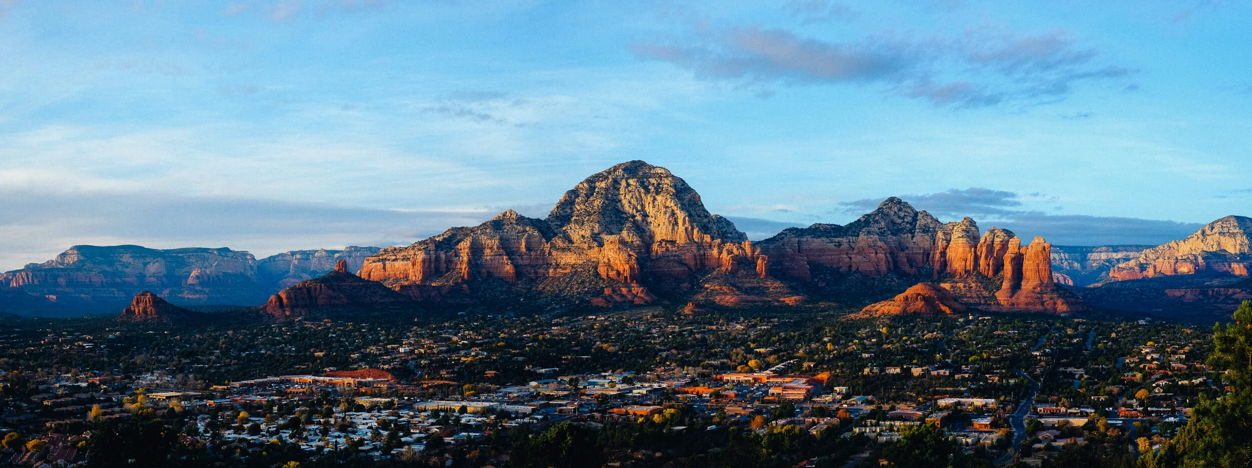 View of the red rock mountains and cityscape in Sedona, Arizona during sunset with blue sky and scattered clouds.