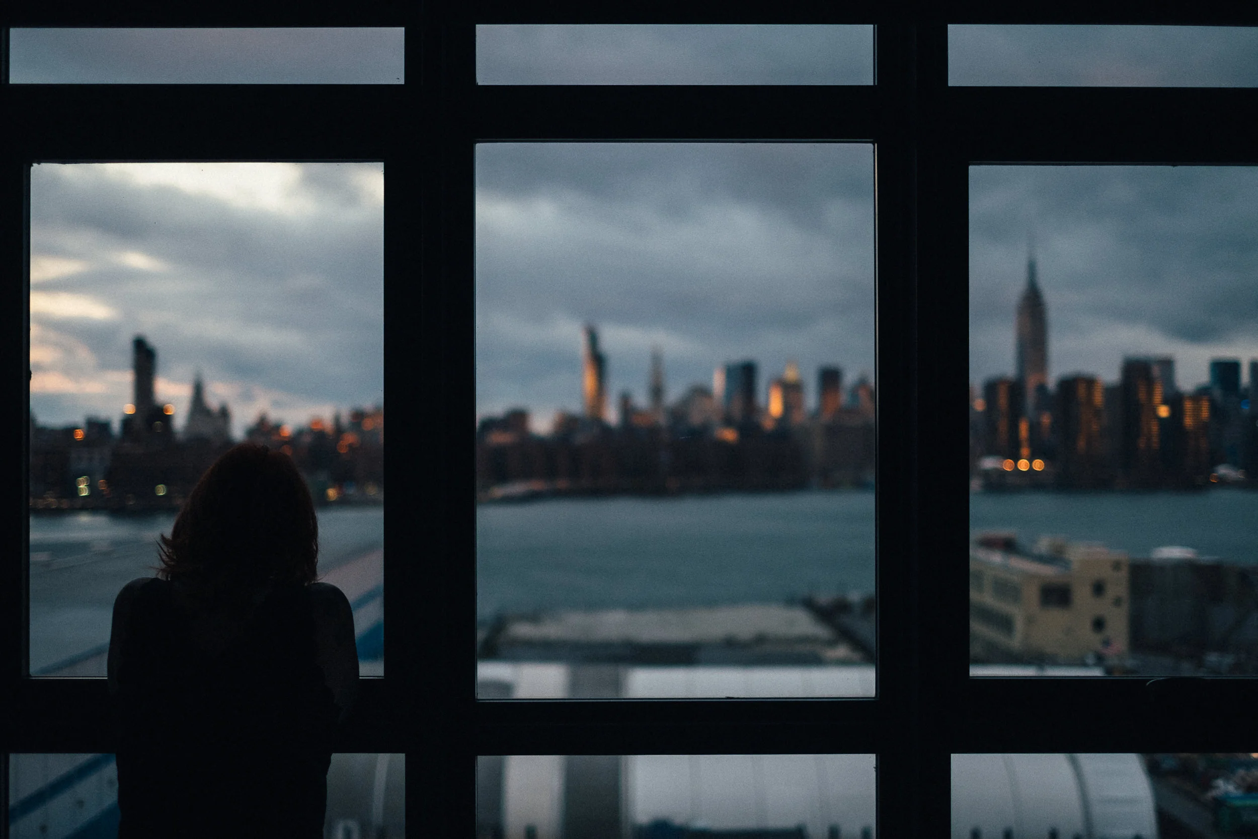 A silhouette of a person with shoulder-length hair looking out a large window at a city skyline with tall buildings and a cloudy sky during dusk.