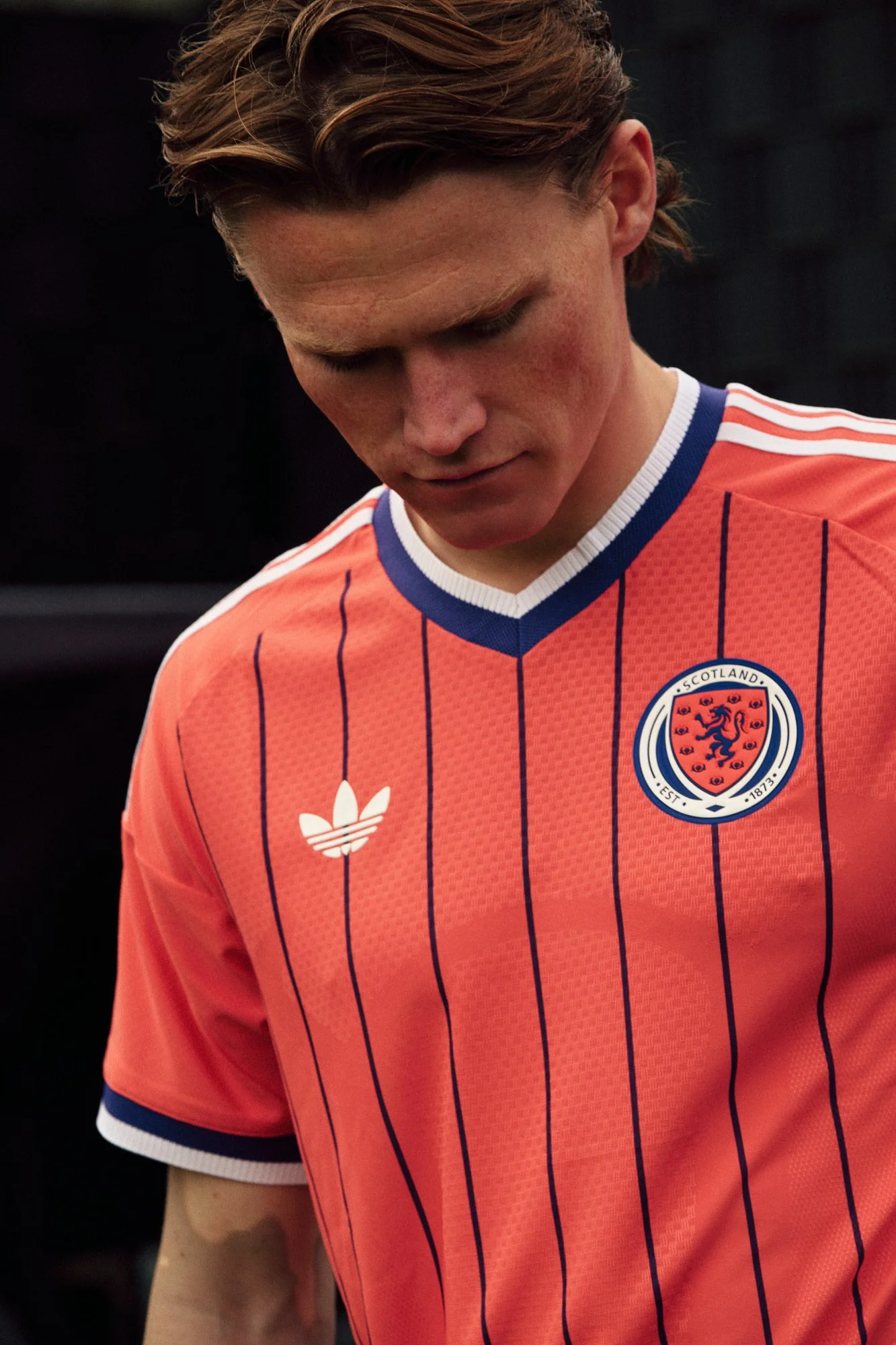 Close-up of a person wearing a Scotland national soccer team jersey, looking downward. The jersey is orange with blue pinstripes, and features the Scotland crest and Adidas logo.