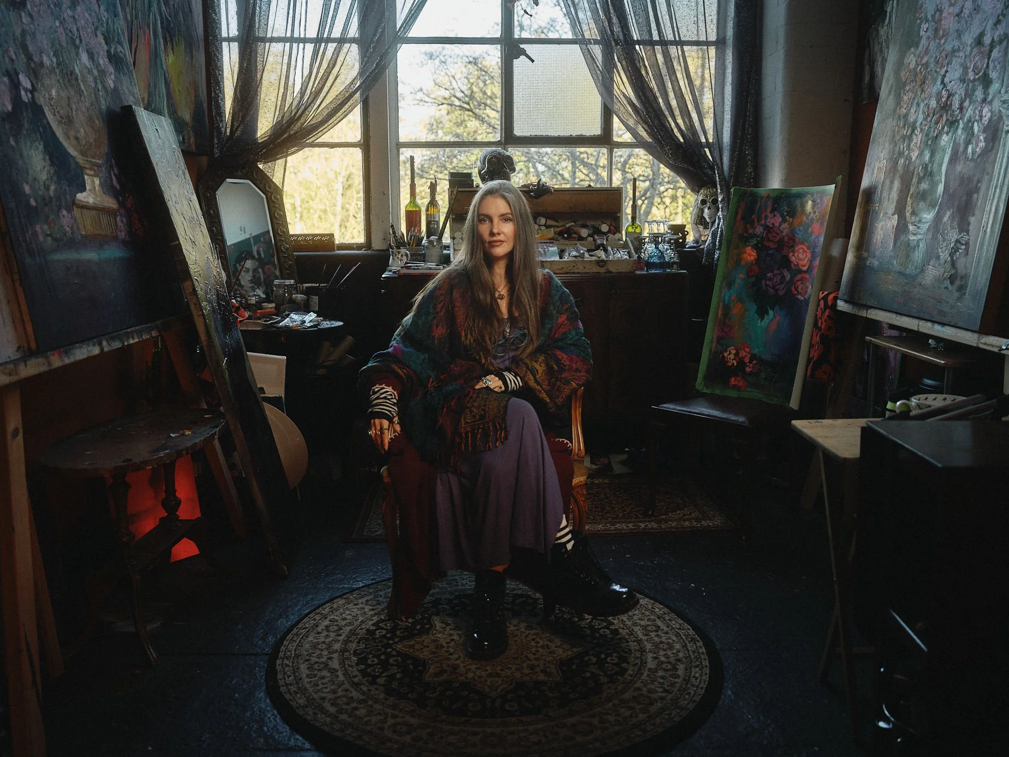 A woman with long hair sitting inside an artist's studio surrounded by paintings, art supplies, and sculptures, with sunlight coming through large windows behind her.