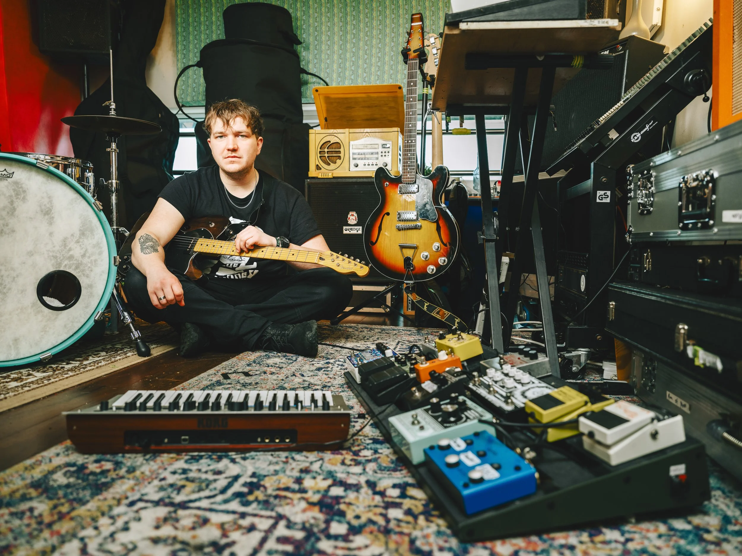 A young man with brown hair holding an electric guitar in a music studio filled with instruments and equipment, including drums, keyboards, effects pedals, and amplifiers.