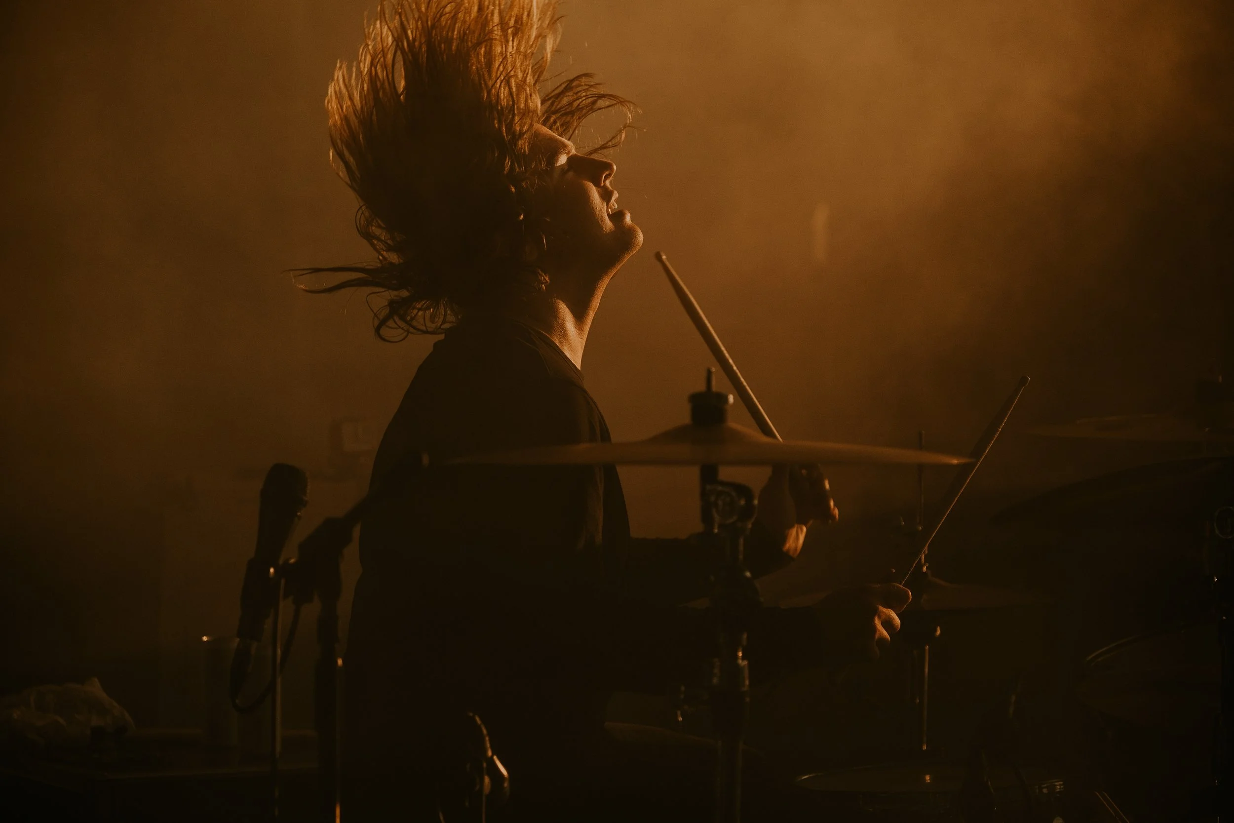 A drummer with long hair is playing drums on stage, illuminated by warm lighting.
