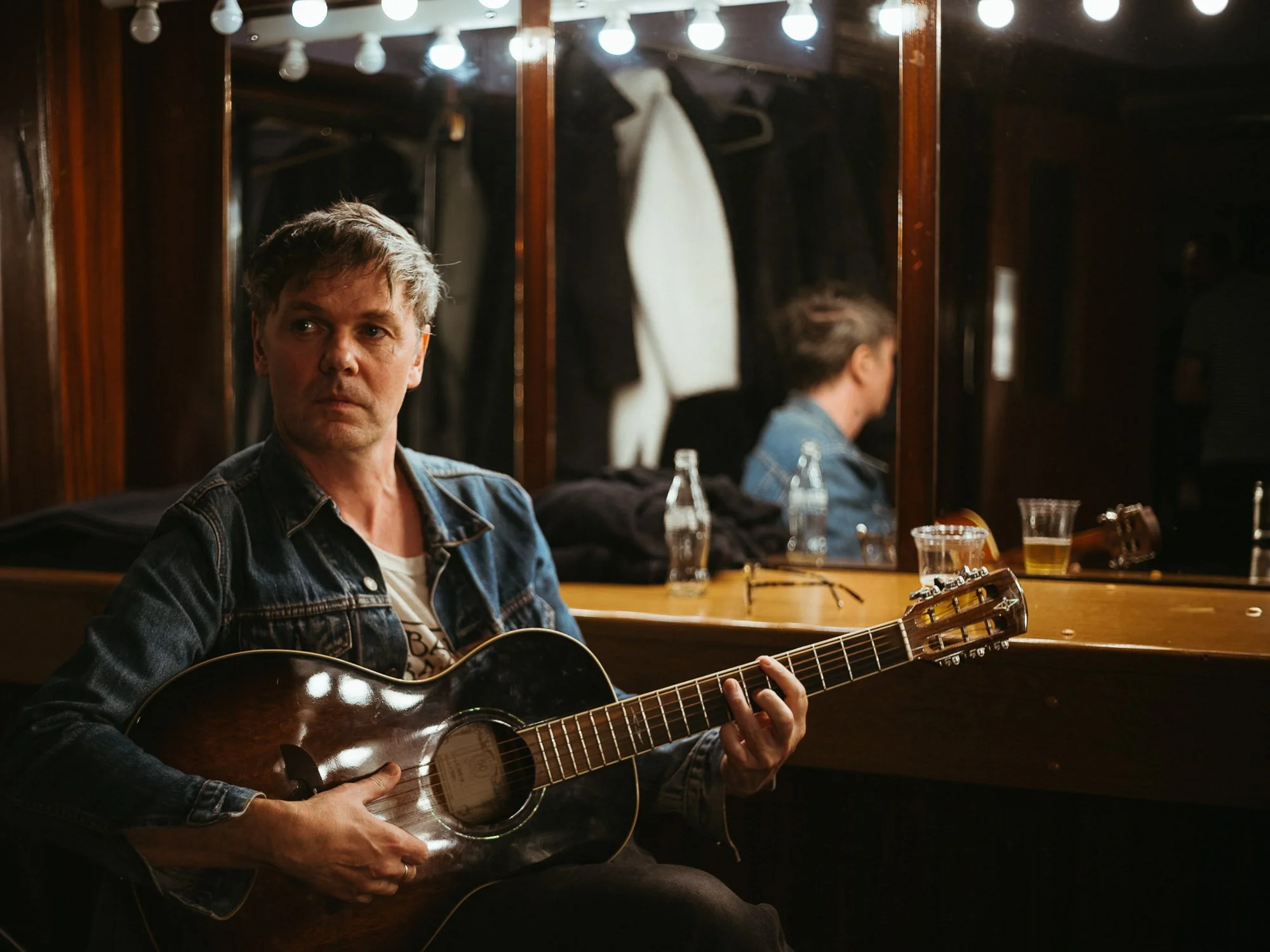A man with short, grayish-brown hair playing an acoustic guitar in front of a mirror in a dimly lit room with string lights.