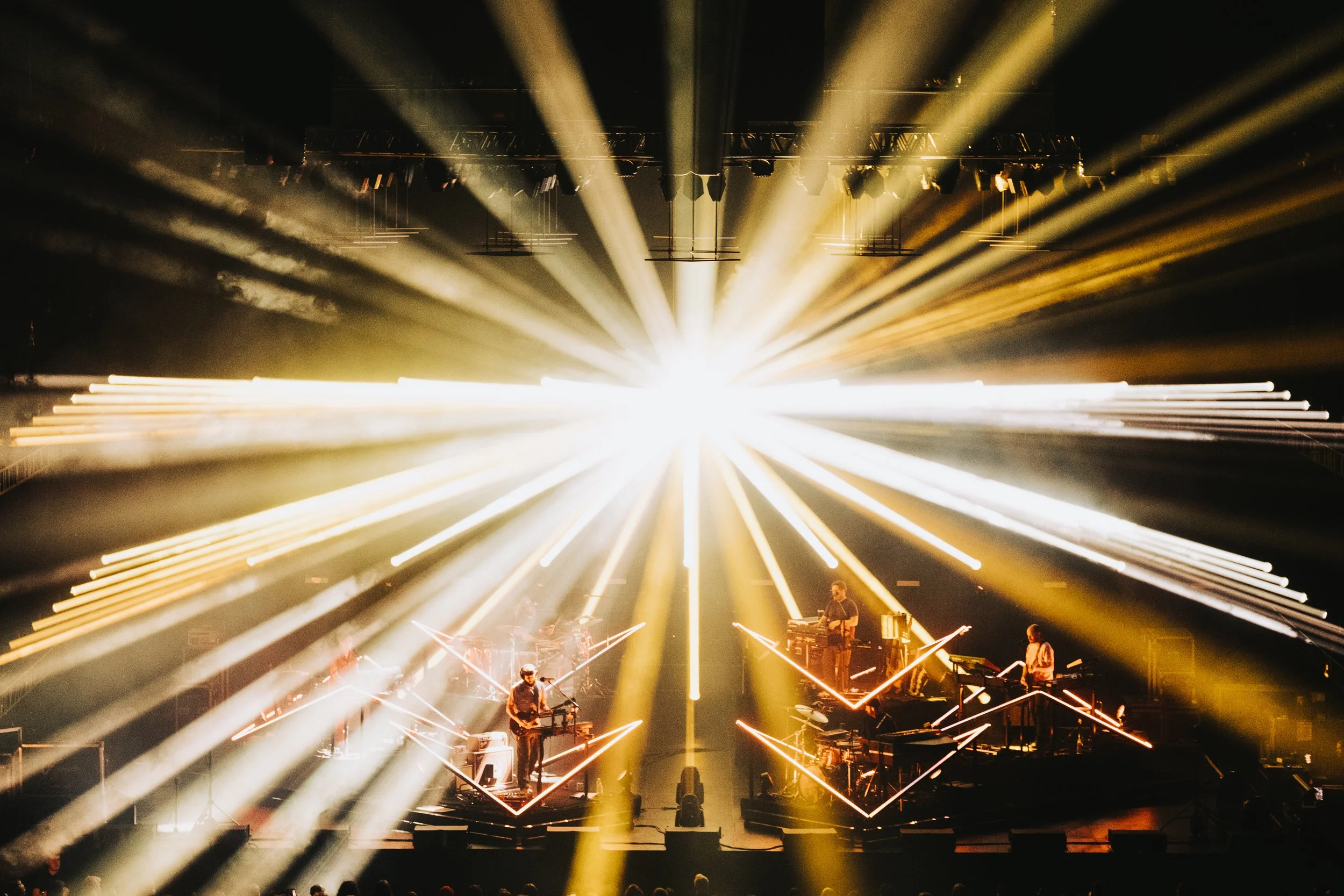 Musicians performing on stage at a concert with dramatic yellow and white stage lighting and light beams radiating outward.